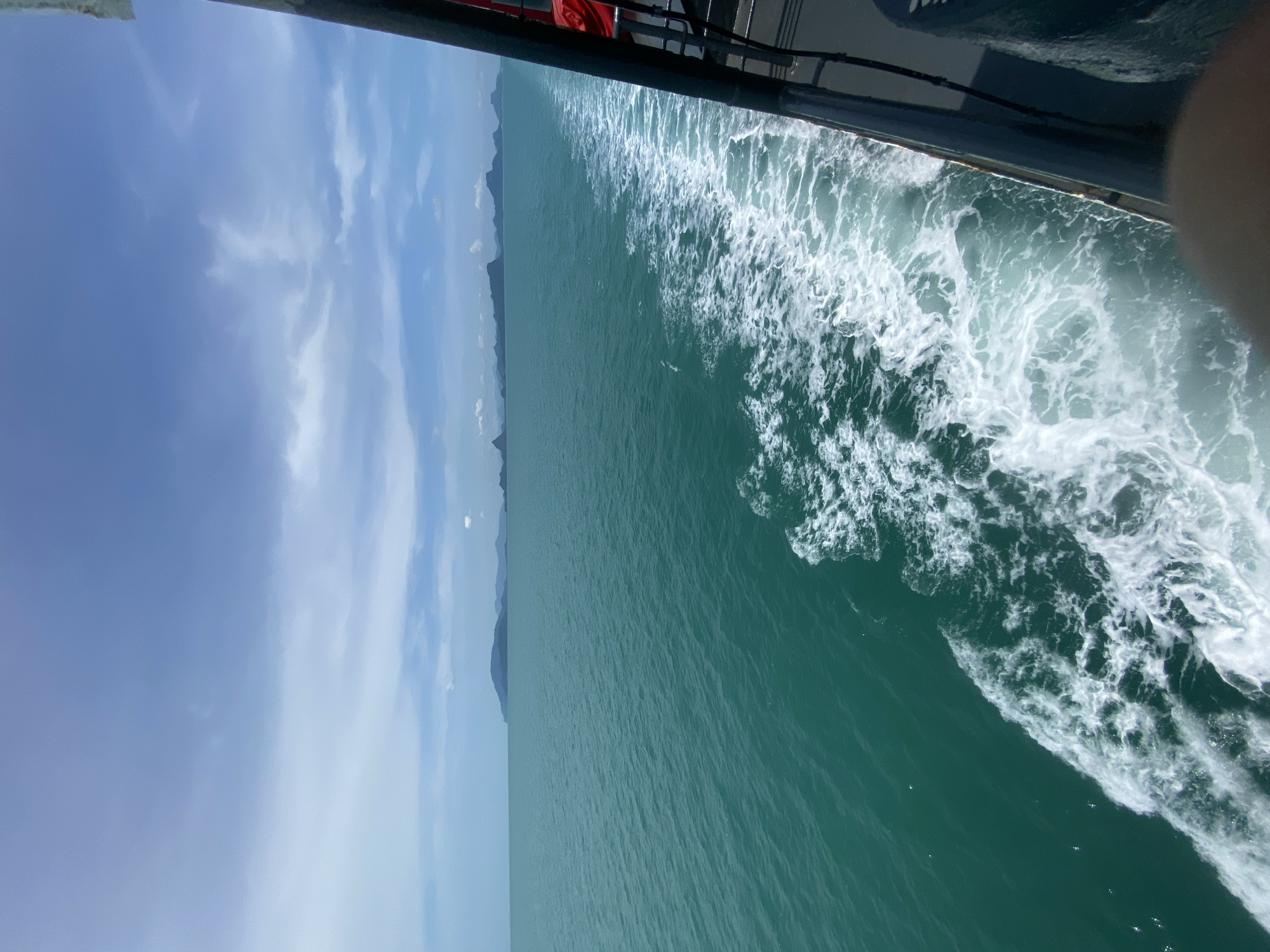 The image depicts a view from the side of a moving ferry boat on a clear day in the waters off Thong Nian, Thailand. The sea is a vibrant turquoise, with a white wake trailing behind the vessel, indicating forward motion. In the distance, a chain of islands with mountainous terrain is visible against a pale blue sky dotted with wispy clouds. The right side of the frame shows a portion of the ferry's railing and some dark superstructure, with a hint of a red life vest. The overall scene conveys a sense of travel and the natural beauty of the tropical seascape.