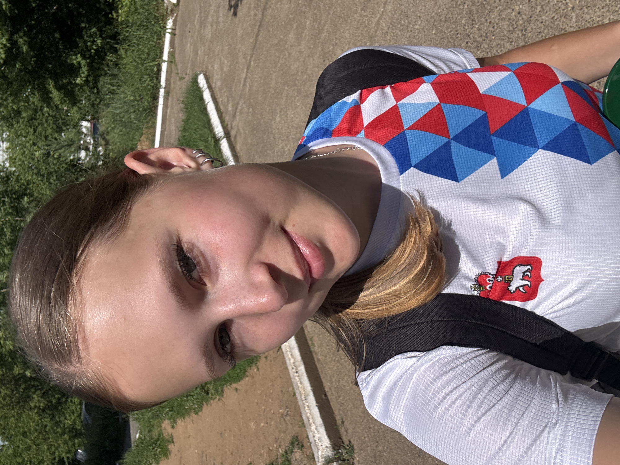 This is a close-up, outdoor selfie of a young Caucasian woman with blonde hair, taken during the daytime in Chaikovsky, Russia. She is wearing a white t-shirt with a red, white, and blue geometric pattern on the right side of the chest, and a red crest with a white bear and crown emblem on the left side. She is also wearing a black backpack. The background shows a paved path, a grassy area, and trees. The scene appears to be a public park or recreational area.