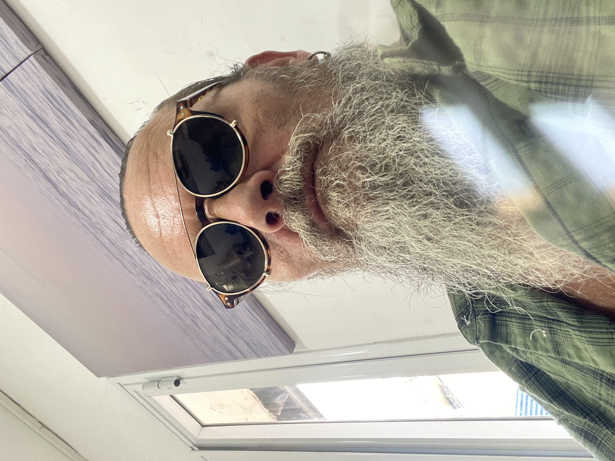A close-up, low-angle, selfie-style photograph captures a man from the chest up, indoors. He has a long, gray beard and mustache, and wears round, tortoiseshell-rimmed sunglasses with dark lenses. His head is bald on top, with some gray hair around the sides. He is wearing a green plaid shirt. To the left of the frame, a white window with vertical panes is visible, with what appears to be a building outside. The background consists of a white wall and a large mural or print of wavy water. The man is positioned centrally in the frame, with the camera focused on his face and upper torso. The lighting is bright, suggesting daylight. The scene appears to be in an interior space, potentially a room or office, and the presence of the mural and window suggests an urban setting. The man's gaze is directed towards the camera. There is no indication of any specific location in Netanya, Israel within the visual information provided.