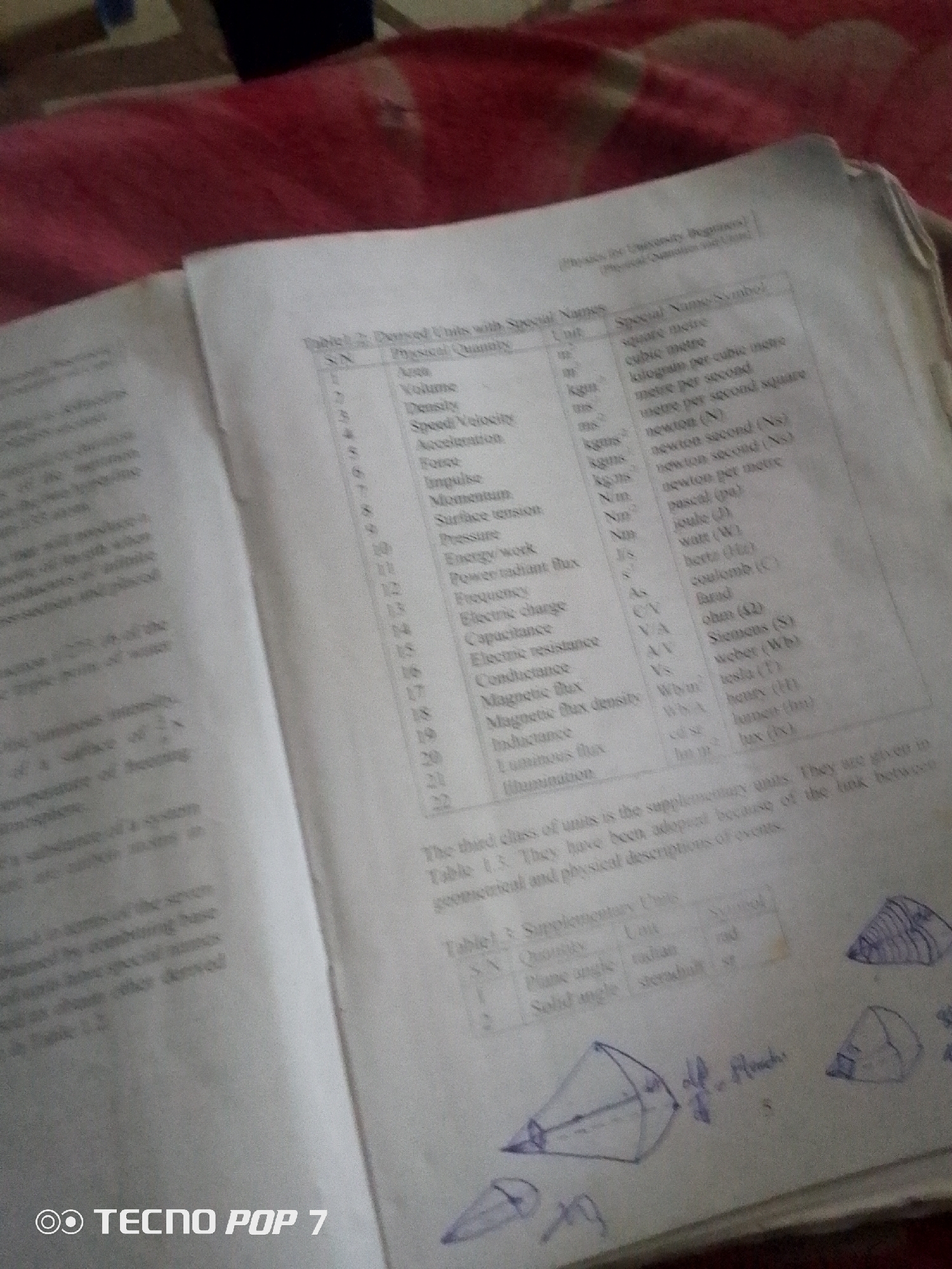 This media file contains a close-up shot of an open book lying on a red, textured surface, possibly a bed or blanket. The book appears to be a textbook, with visible tables of scientific units and their corresponding symbols and names. There are also handwritten annotations and sketches of geometric shapes, such as cones, on the pages.
The text in the book includes titles like "Derived Units with Special Names" and "Supplementary Units," along with columns for SN, Physical Quantity, Unit, and Special Name/Symbol. Specific units mentioned include square meter, cubic meter, newton, joule, watt, ohm, and weber. There are also handwritten notes like "of all which".
The scene is indoors, with no clear indication of the time of day or weather conditions. The location is suggested to be Ipetumodu, Nigeria, based on external context. The overall impression is one of a study or learning environment, with someone actively engaging with the material through reading and note-taking. The presence of the "TECNO POP 7" watermark in the lower corner indicates the image was taken with a Tecno Pop 7 smartphone.