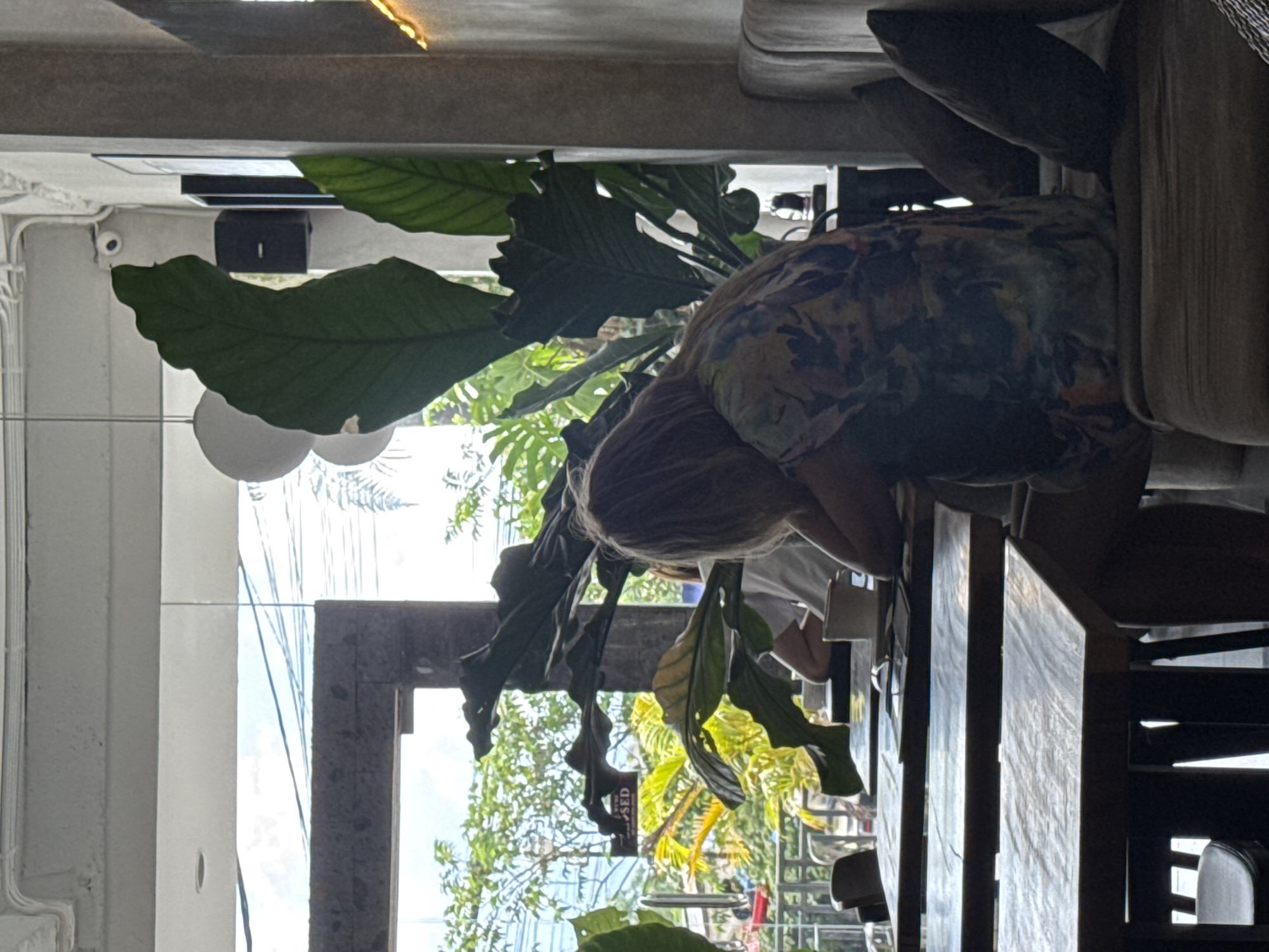 An indoor scene, likely a cafe or restaurant in Kuta Selatan, Indonesia, shows a person seated at a wooden table, facing away from the camera. The person has light brown hair and is wearing a patterned or floral-print dress or shirt, with their head bowed or resting on their hand, suggesting relaxation or concentration. Another person's arm and a white shirt are partially visible on the right, also seated at the table, with a white cup nearby.
The setting is characterized by a blend of natural and modern elements. Large-leafed tropical plants, possibly Monstera, are prominently placed in the foreground and midground, creating a lush, green ambiance. Behind the seated individuals, a large window or open section reveals bright daylight, sunny weather, and dense green foliage of trees outside, typical of a tropical environment.
Architectural details include a concrete ceiling, white walls, and dark window or door frames. A small white security camera is mounted near the ceiling on the left, with a black speaker or electronic device below it. Two spherical white lamps hang in the midground, partially obscured by leaves, and a string of decorative lights runs along the ceiling on the far left.
Outside the window, a partially visible sign reads "CLOSED," hanging from what appears to be an exterior door or gate. The overall atmosphere is bright and tropical, consistent with its location.