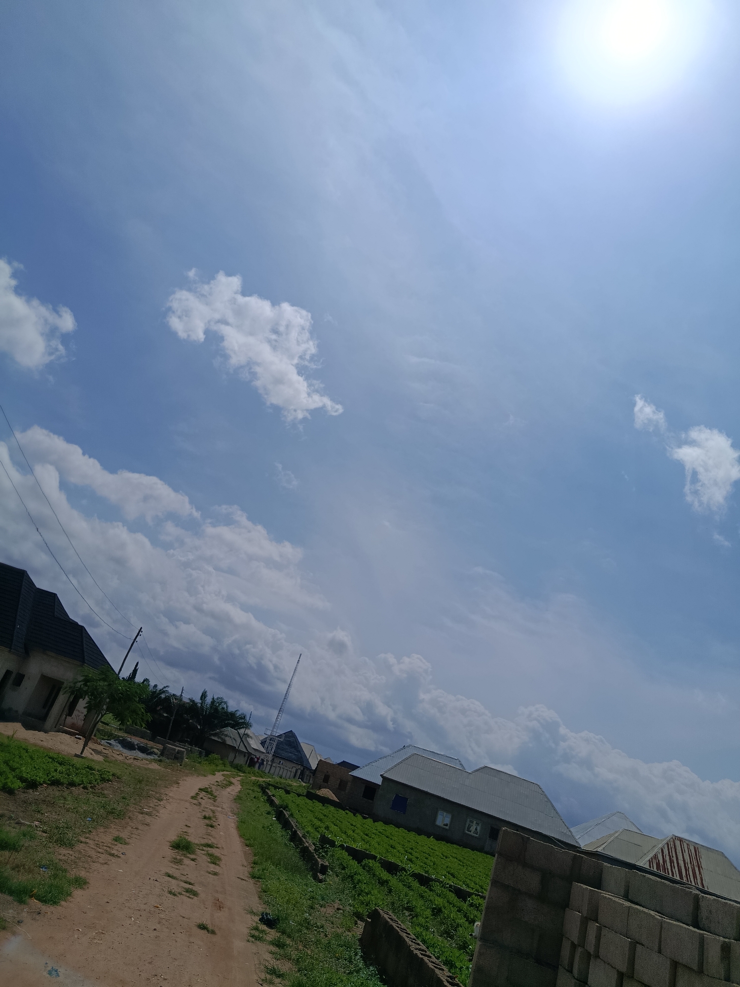 The image shows a sunny day in New Nyanya, Nigeria, with a bright sun in the upper right corner of the frame. The sky is predominantly blue with scattered white clouds. In the foreground, a dirt road winds through a residential area with several houses visible. Some of the houses are under construction, with visible concrete blocks and unfinished roofing. Lush green vegetation, likely crops or gardens, lines the sides of the road. A few trees are also present, one of which is growing next to a house on the left. In the background, a tall telecommunications tower is visible. The overall scene depicts a developing urban or semi-urban environment with evidence of ongoing construction and agricultural activity. There are no people visible in the image.