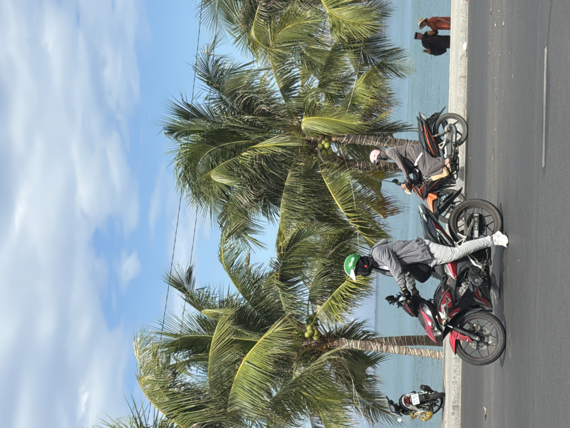 The image depicts a coastal scene in Nha Trang, Vietnam, characterized by a road in the foreground, palm trees, and the ocean in the background. Two individuals are riding motorcycles on the road. The first motorcyclist, in the foreground, is wearing a gray jacket, gray pants, a green helmet, and a black face covering. Their motorcycle is red and black. Further back, a second motorcyclist is wearing a gray outfit and a pink head covering, riding an orange and black motorcycle. To the right, two individuals are standing near the ocean, one in a dark outfit and the other in an orange dress with a straw hat. The ocean is a vibrant blue, and the sky is clear with some scattered clouds. Power lines are visible between the palm trees and the sky. A parked motorcycle is visible on the left side of the image.