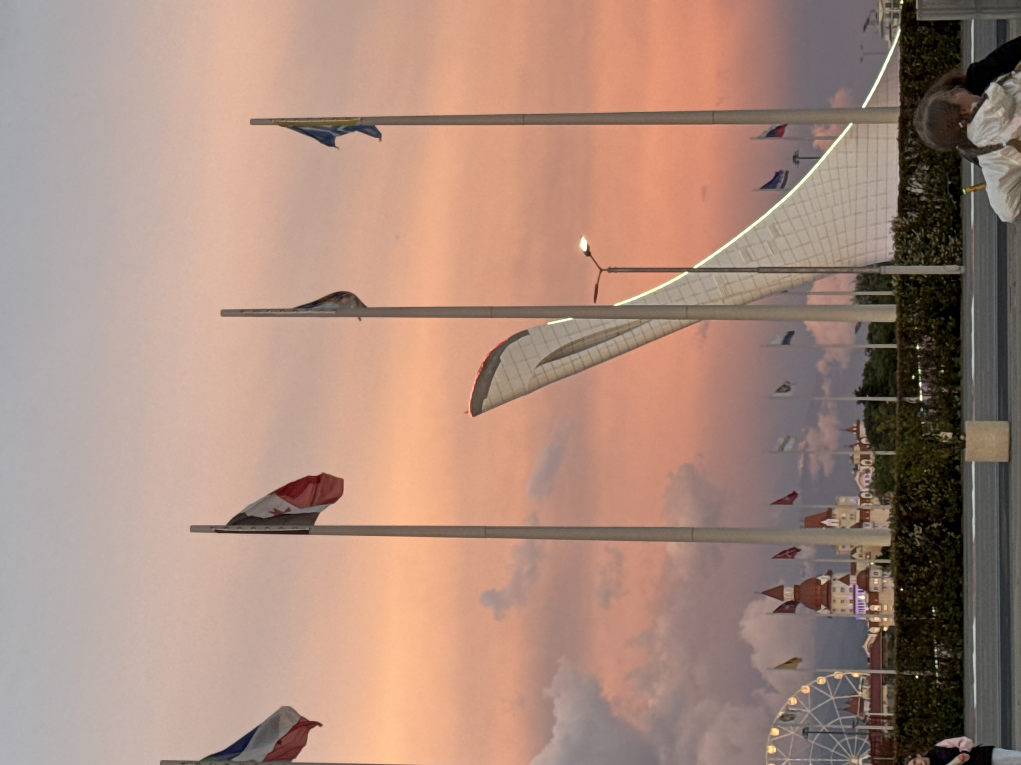 The image, taken in what appears to be Sirius, Russia, shows a sunset sky with a variety of flags mounted on flagpoles. The flags appear to be national flags of different countries. Some of the flags are partially obscured or difficult to identify due to being limp. There is a large Ferris wheel and buildings, some styled in a medieval fashion, suggesting an amusement park or themed area. A large, white, arching structure, perhaps part of a stadium or architectural design, is also visible. On the right, there is a paved area with what looks like a hedge. A person in white clothing is visible in the upper right corner. The overall scene suggests a public gathering place with international themes, possibly a festival or exhibition. The setting sun gives a warm, peach-toned hue to the sky.