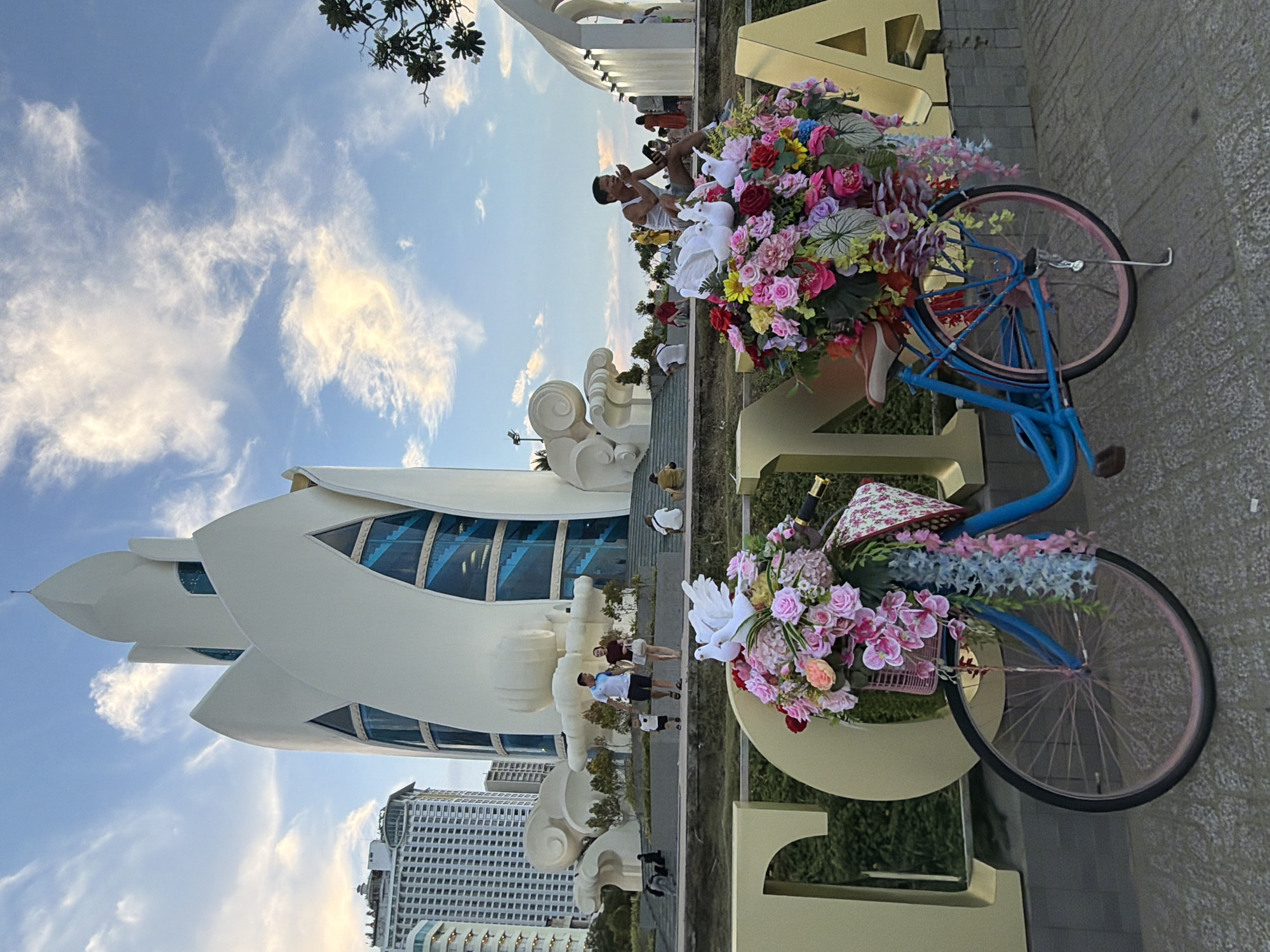 In Nha Trang Ward, Vietnam, a decorative blue bicycle laden with vibrant floral arrangements, including pink, red, and purple roses and hanging greenery, stands prominently in the foreground. Two white dove figures are perched atop the floral display. Behind the bicycle, large golden letters form part of a sign, with "T", "C", "N", and "A" clearly visible. In the middle ground, several people are visible, including a family walking with a child and a man sitting on a ledge. The backdrop features a modern, white, lotus-shaped building with blue windows, and several high-rise buildings. The sky is a clear blue with scattered white clouds, suggesting late afternoon or early evening light.