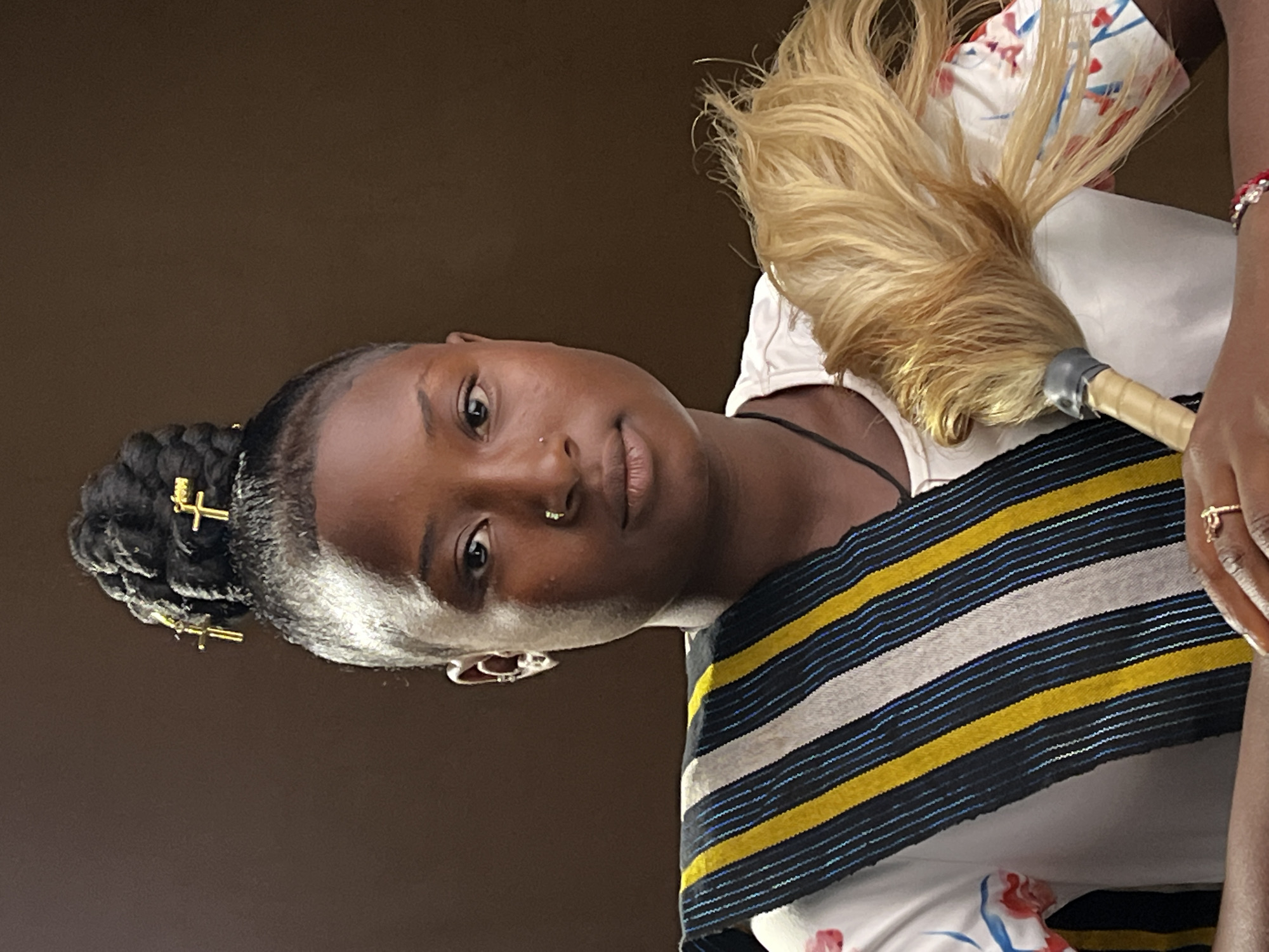 A head-and-shoulders portrait features a young person, likely a woman, with dark skin, looking directly at the viewer. Her dark hair is styled in neat braids, pulled back into a bun on the left side of her head (viewer's left), adorned with two small, gold-toned cross-shaped hair accessories. She has a small stud nose piercing on her right nostril and a distinctive silver ear cuff on her left ear. Her expression is calm and neutral, with a direct gaze.
She is wearing a white short-sleeved top, with faint floral patterns in red and blue visible on the sleeves and the lower portion of the garment. Over this, she dons a horizontally striped garment or stole, featuring prominent bands of black, yellow, and white, with thin, shimmering blue lines woven into the black sections.
With her right hand, visible on the right side of the frame, she holds a whisk-like object. This object has a light-colored handle, possibly wood, secured with silver tape where it meets a voluminous, blonde or light-colored hair-like top section. Her right wrist is adorned with a bracelet featuring red and clear beads, and a gold ring with a small, looping design is visible on her finger. The background is a plain, solid dark brown, providing a stark contrast to the subject. The setting appears to be indoors or against a simple backdrop, likely in Jos, Nigeria. No visible text is present.
