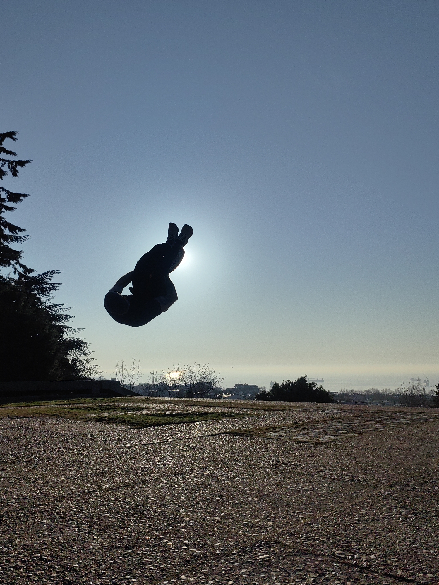 The image displays an outdoor scene in Varna, Bulgaria, captured from an elevated viewpoint overlooking the city and the Black Sea. A solitary individual is depicted mid-air, silhouetted against the sun, executing an acrobatic flip with their body tucked into a compact form, knees drawn towards the chest. The sun, positioned behind and slightly above the person, casts a strong backlight, rendering the individual and nearby vegetation in dark silhouette against the clear, light blue sky.
The foreground consists of a textured ground surface, appearing to be paved with exposed aggregate concrete or small pebbles, interspersed with patches of sparse green grass. To the left, a large, dark coniferous tree, possibly a pine or cypress, is partially visible, also silhouetted. In the midground and background, the urban landscape of Varna spreads out, featuring various buildings and bare deciduous trees. Beyond the city, the vast expanse of the Black Sea is visible, appearing somewhat hazy. Several large cargo ships are discernible on the water, and the sun's reflection creates a bright glint on the sea surface. This panoramic perspective suggests a location within or adjacent to Varna's Sea Garden (Morska Gradina) or a similar elevated viewpoint overlooking the port and bay.