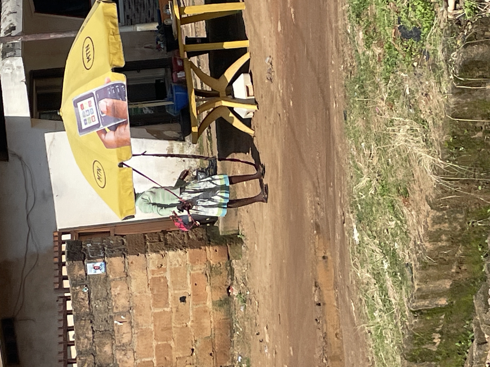 In Jos, Nigeria, a person stands under a yellow umbrella emblazoned with the MTN logo and an image of a hand holding a mobile phone. The person is wearing a light green long-sleeved shirt and a patterned skirt. They are barefoot and holding a red string. To the right of the umbrella is a yellow table with some items on it, including a blue basin. Behind the person and the umbrella is a brick wall. The ground is dirt and there is some overgrown grass and debris in the foreground and background.