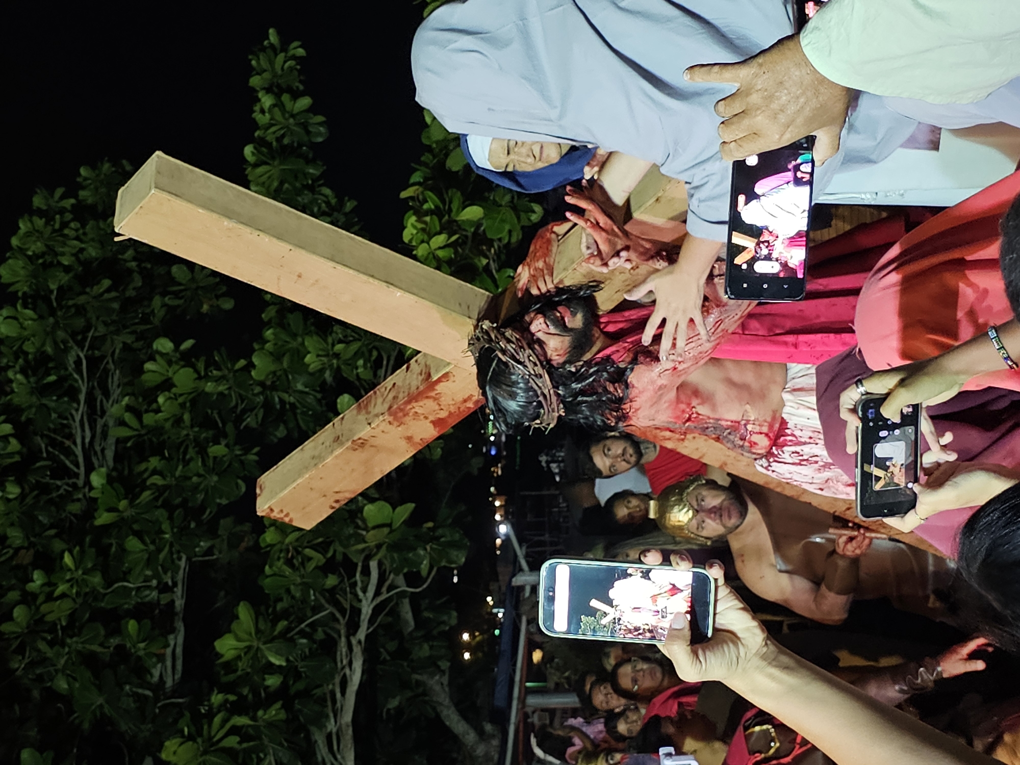 A nighttime scene in Armação dos Búzios, Brazil, shows a reenactment of the crucifixion of Jesus Christ. A man portraying Jesus, with blood and a crown of thorns, is affixed to a wooden cross. The cross is positioned in front of a leafy tree. A woman in a nun's habit stands near the cross, along with others surrounding the reenactment. Several onlookers are holding smartphones, apparently taking photos or videos of the scene.