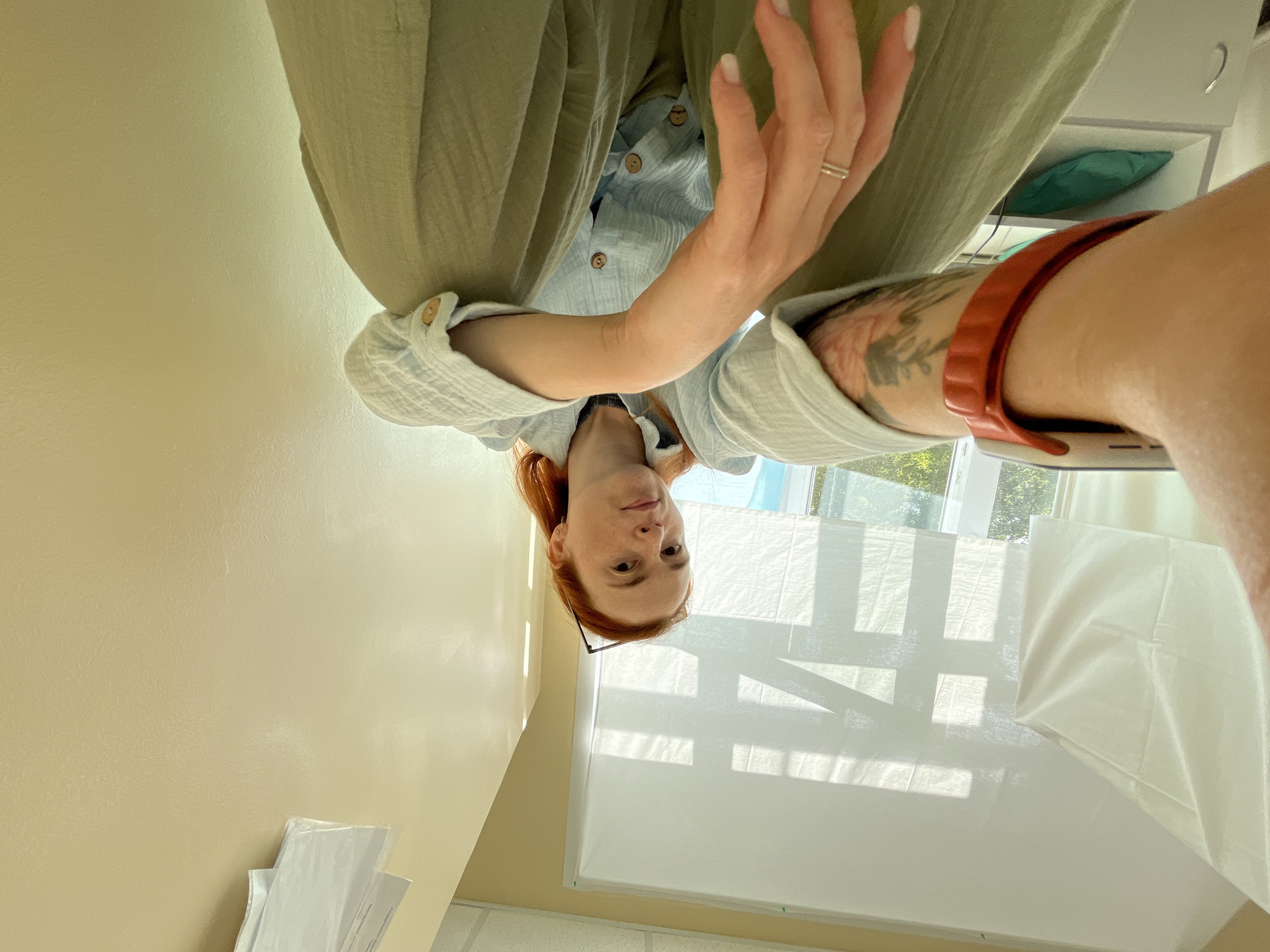 A woman with red hair is sitting on the floor and taking a selfie. She is wearing a light blue gauze shirt and green pants. She has a red fitness tracker on her left wrist and a ring on her right ring finger. The background is a room with a yellow wall and a window with a white blind. There are some papers tacked to the wall above the blind.