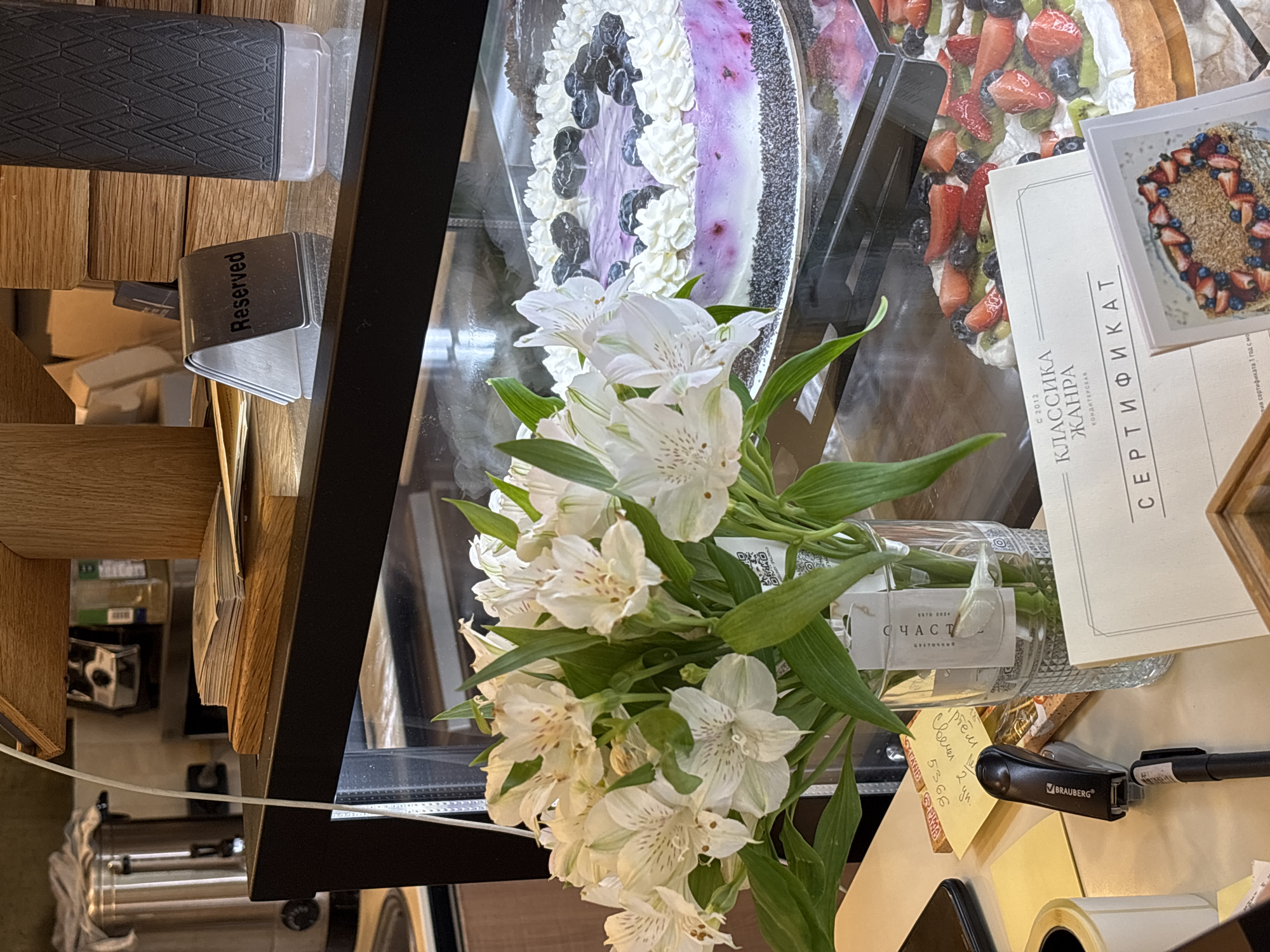 This is an interior shot, likely taken inside a pastry shop or cafe, potentially in Perm, Russia.
The scene is dominated by a display case containing cakes. One cake features a blueberry topping and whipped cream, while another is decorated with strawberries, blueberries, and kiwi. There's a promotional card near the cakes, along with a certification document written in Russian.
A bouquet of white Alstroemeria flowers in a glass vase is positioned in front of the display case. A "Reserved" sign is visible, suggesting a table or area is not available. A label on the vase reads "СЧАСТЬ" (Happiness).
Other items present include a sticky note with handwritten text, a stapler branded "BRAUBERG", a roll of tape, and a phone or electronic device.