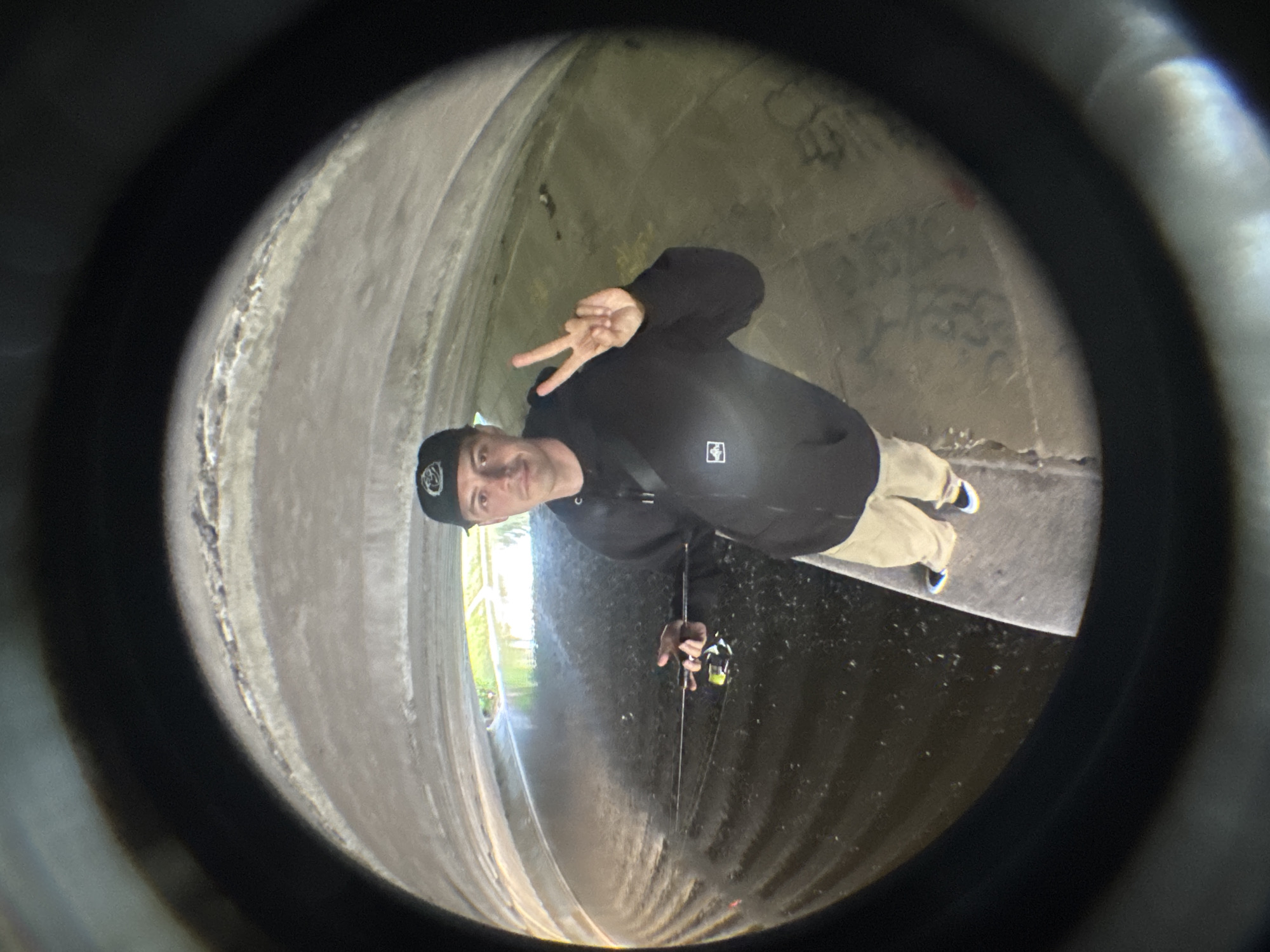 A man wearing a black hoodie, tan pants, and a black baseball cap stands in a concrete tunnel with a fishing rod. He is making a peace sign with his right hand. The tunnel appears to be a storm drain or sewer. The water in the tunnel is dark and murky, and there is some graffiti on the walls. The image is taken with a fisheye lens, which distorts the perspective and makes the edges of the image appear warped and dark. The man is the main subject of the image, and he is in focus. The background is blurred and out of focus. The lighting in the tunnel is dim and artificial. There are no other people or animals visible in the image. The overall mood of the image is mysterious and slightly unsettling. The location is not specified in Saint Petersburg, Russia.
