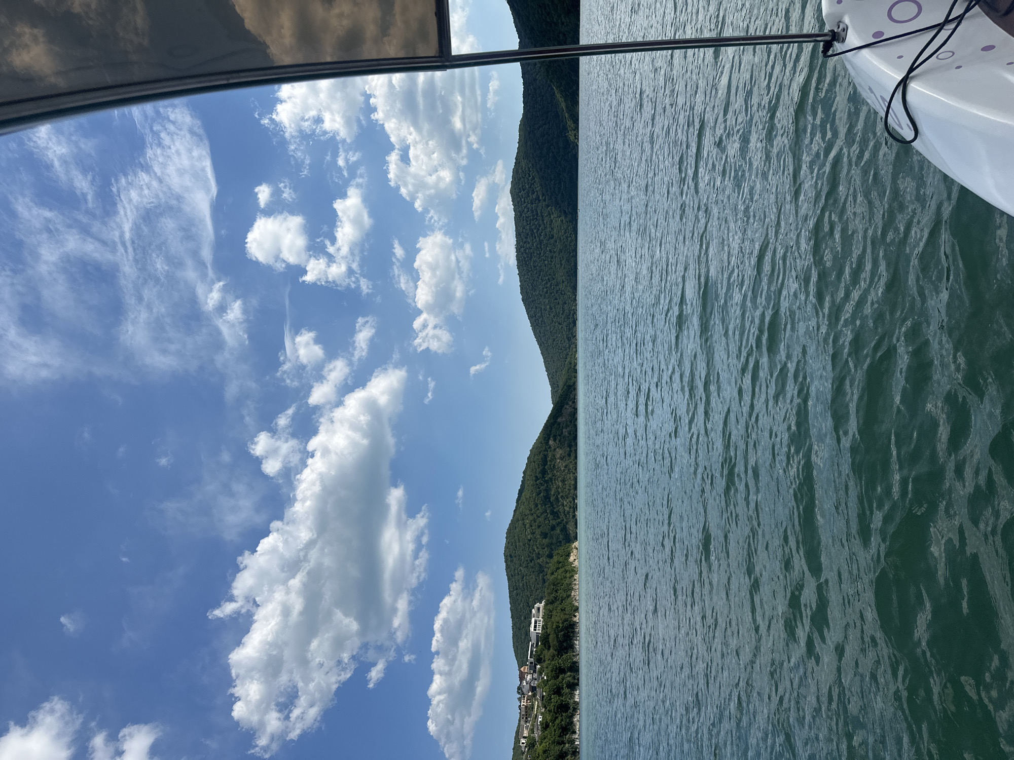 The image depicts a view from a boat on the Abrau Lake in Abrau-Dyurso, Russia. The lake's water is a deep blue-green, with small waves rippling across its surface. In the distance, lush green hills covered in dense vegetation rise from the water's edge. A small settlement with houses and buildings is visible on the slope of one of the hills. The sky above is a vibrant blue, adorned with scattered white cumulus clouds. The right side of the image shows a portion of the boat, including a white hull with purple circular patterns and a metal railing. The overall scene conveys a sense of a peaceful day on the water, surrounded by natural beauty.