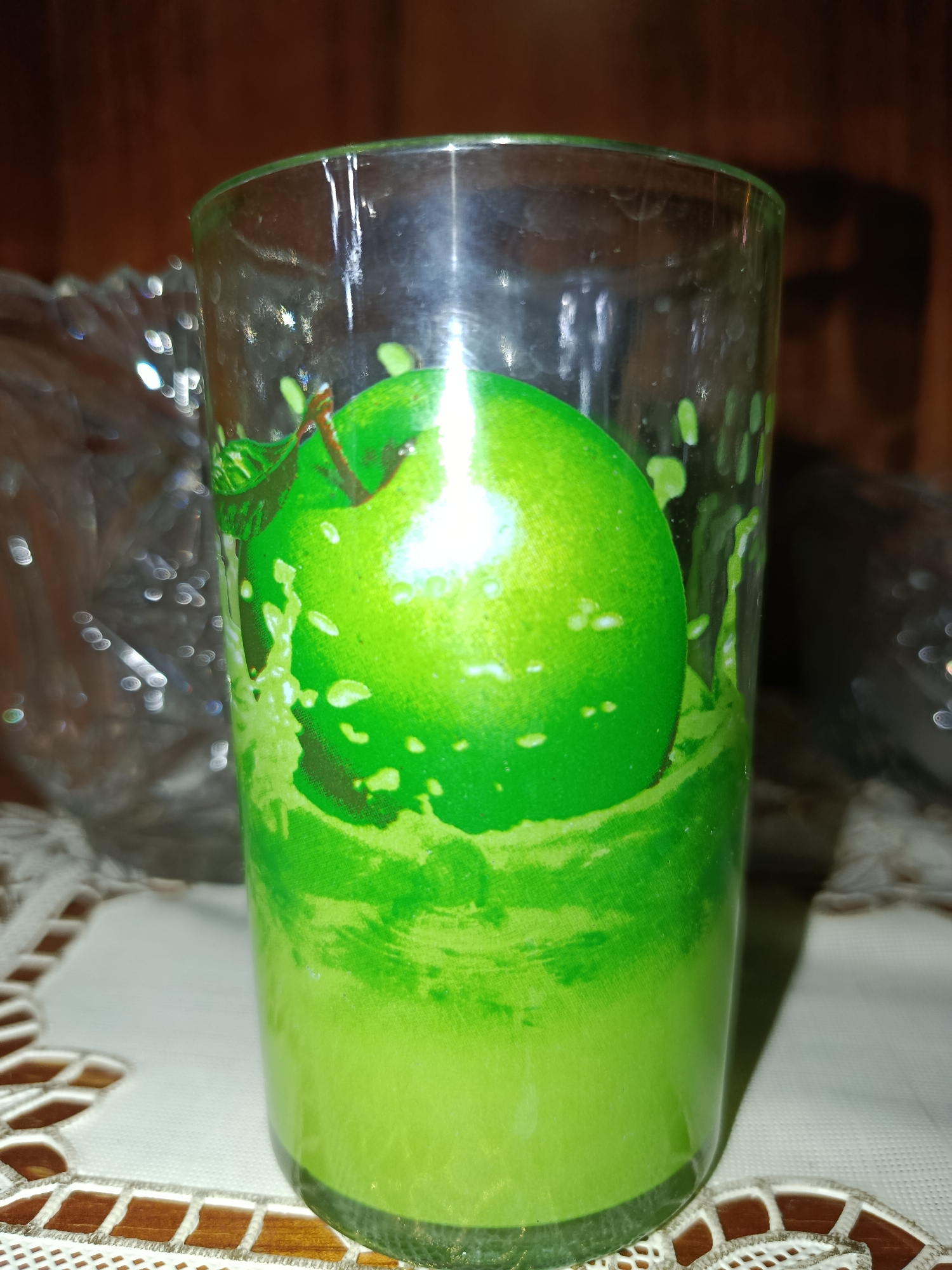A tall, clear glass with a green apple design and a green liquid inside sits on a white crocheted placemat. The background is dark and out of focus, with some indistinct shapes and lights visible. The image appears to be taken indoors, likely in a home setting. There is no indication of a specific location in Tashkent, Uzbekistan.