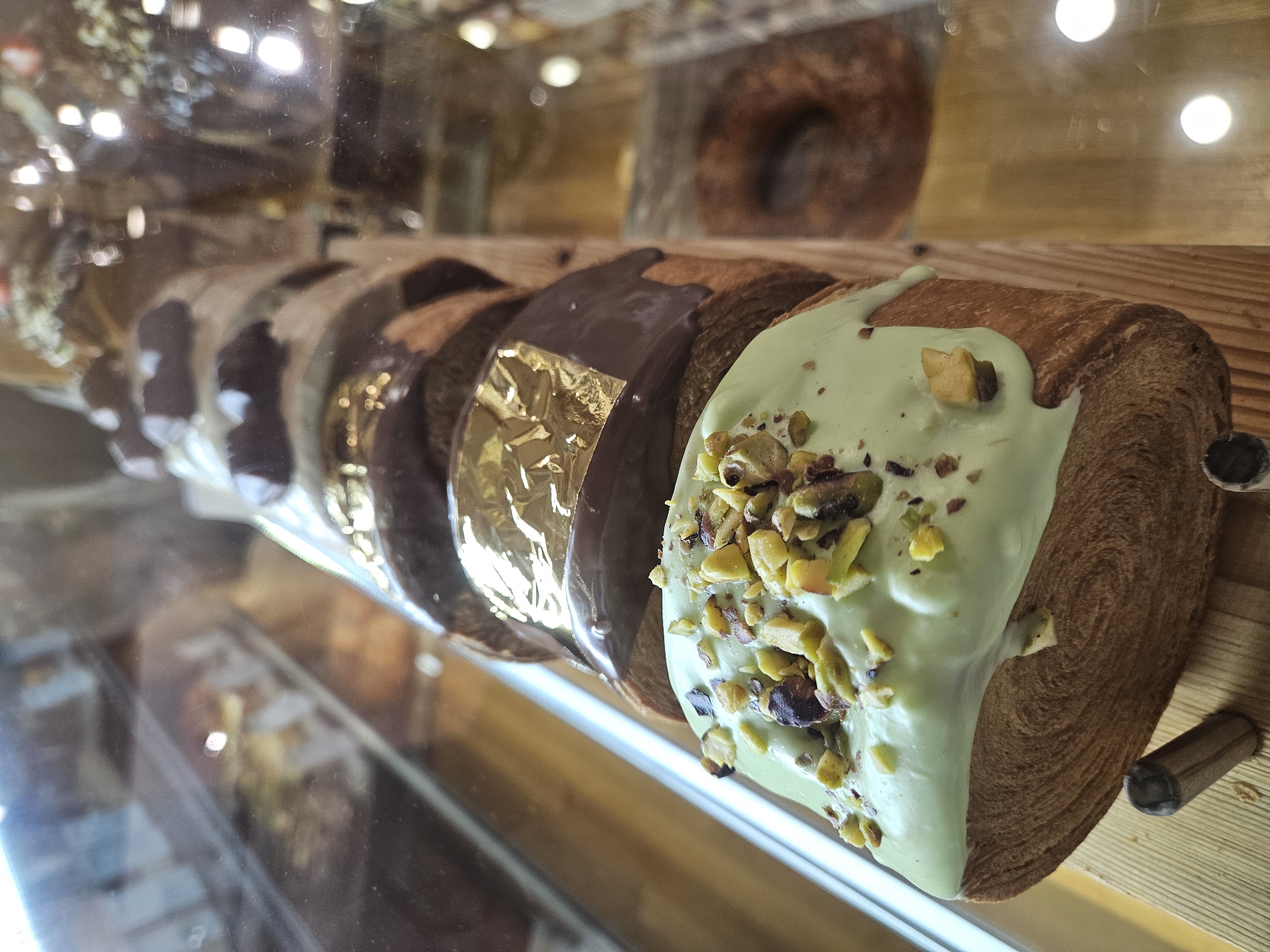 The image displays a close-up, eye-level view of a row of pastries arranged diagonally within a glass display case, likely in a bakery or cafe setting. The main subjects are several long, cylindrical pastries, characteristic of a kronut or laminated dough pastry, resting on a light brown wooden surface.
In the foreground, the most prominent pastry is topped with a vibrant, light green glaze that appears to drip slightly down its sides. This green glaze is heavily garnished with chopped nuts, primarily pistachios, which add a contrasting texture and color. The flaky, layered structure of the pastry's dough is clearly visible on its exposed side. Behind this pastry, a series of similar pastries extends into the background. Each of these subsequent pastries is coated with a dark, glossy chocolate glaze. One of the chocolate-glazed pastries features a distinctive gold leaf or metallic foil embellishment on its surface.
The background is softly blurred with visible reflections and bokeh lights, suggesting artificial indoor lighting and a clear glass barrier in front of the display. A large, out-of-focus, donut-shaped pastry can be vaguely seen in the upper right background. The scene depicts a static display of confections, presented in an appealing manner. There are no people visible, and no discernible text is present in the image. The lighting suggests an indoor setting, with no indicators for the specific time of day.