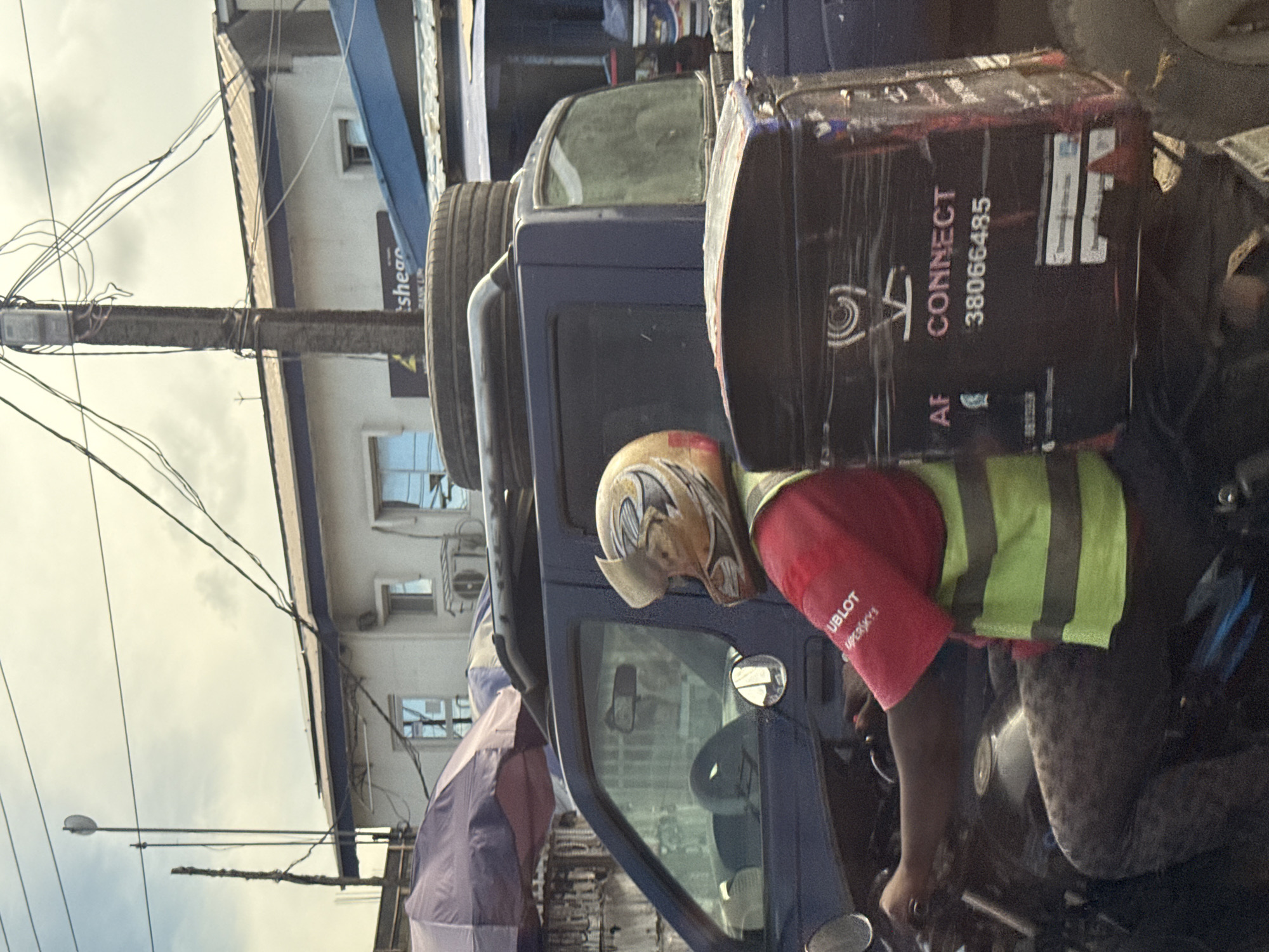 This outdoor image, taken in Ado, Nigeria, captures a daytime street scene under an overcast sky. The central figure is a person seated on a motorcycle or scooter, wearing a light-colored helmet with a swirling design, a red short-sleeved shirt, and a bright green reflective vest with vertical stripes. A large, dark delivery box is securely mounted on the back of the motorcycle.
Adjacent to the person is a dark blue truck or SUV, with multiple tires stacked on its roof rack. Part of the vehicle's side window and mirror are visible. A purple canopy or umbrella is partially open near the front of this vehicle.
The background features a multi-story building with white walls and blue accents, displaying several windows and external air conditioning units. A prominent utility pole, heavily strung with numerous overhead wires, stands in the foreground, with a street light extending from it.
Visible text details include:
- On the delivery box: "AF CONNECT", a circular logo with a spiral design, and the number "38066485".
- On the red shirt: "HUBLOT" and "Mpersky's".
- On a sign on a building in the background: "tshean" and "BANK L" are partially discernible.
The scene depicts a typical urban environment, likely showing a delivery worker engaged in their daily activities.
