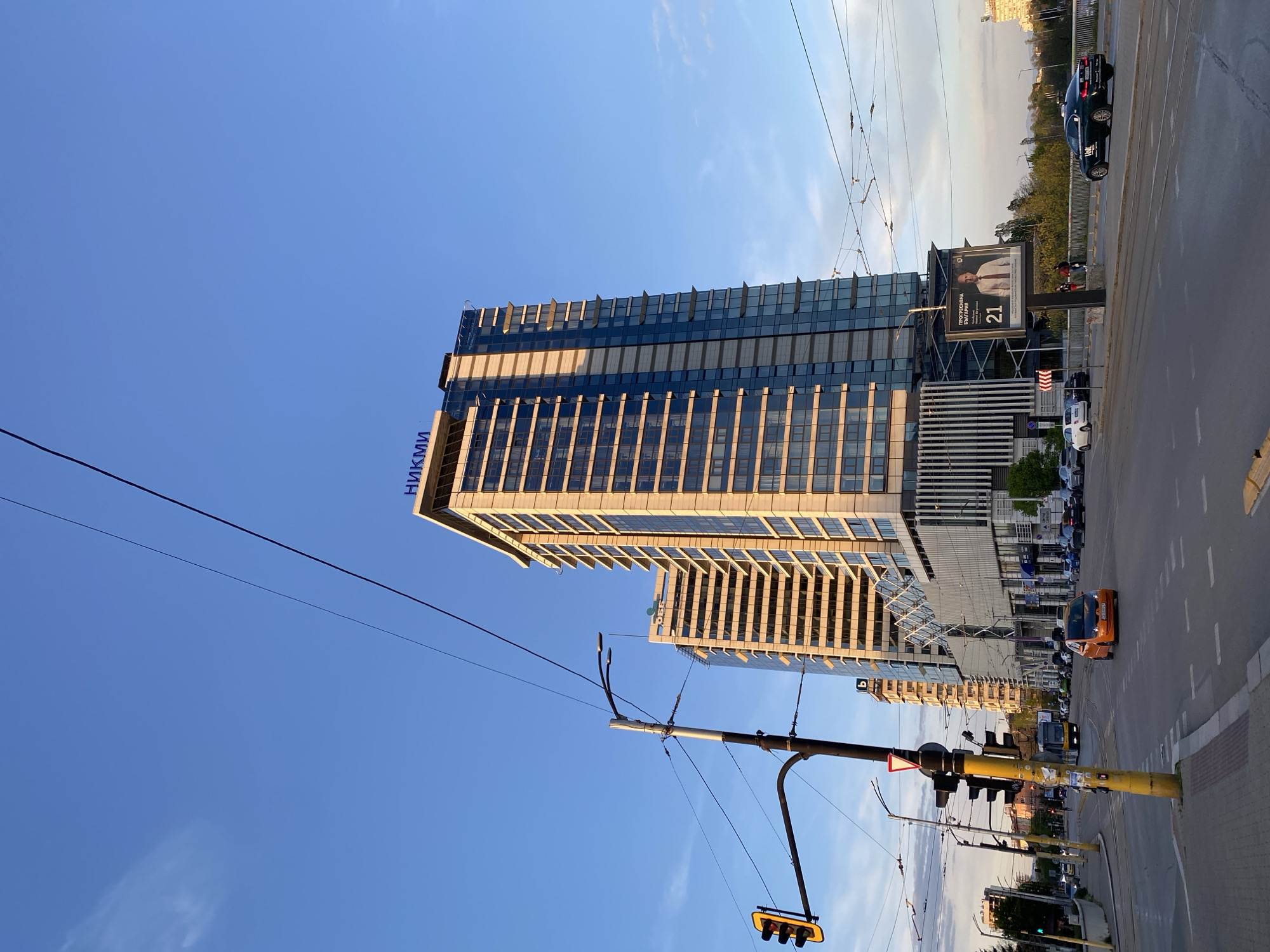 This image depicts a vibrant urban scene in Sofia, Bulgaria, during the late afternoon or early evening, characterized by warm, directional sunlight illuminating the buildings. The sky is clear and blue with minimal cloud cover.
The central subject is a tall, modern high-rise building constructed with a facade of glass and light-toned panels. The Cyrillic text "НИКМИ" (NIKMI) is visible in blue on the upper part of the building. Another, similar building is partially visible behind it. The architecture features prominent vertical lines and a stepped design at the top.
In the foreground and to the right, a multi-lane road is bustling with vehicles, including a distinct orange car and a black sedan. An elevated section of road or a bridge is also visible. A prominent yellow traffic light pole, equipped with several signal heads, stands near the roadside. Overhead, a network of power lines or trolleybus lines crisscrosses the sky.
A large billboard is positioned next to the road, featuring a portrait of a man in a suit. Visible text on the billboard includes "ПРОФЕСИЯ... БИЙДЖЕЙ" (PROFESSION... BIJAY) and the number "21". The scene captures a dynamic city environment with contemporary architecture and infrastructure.