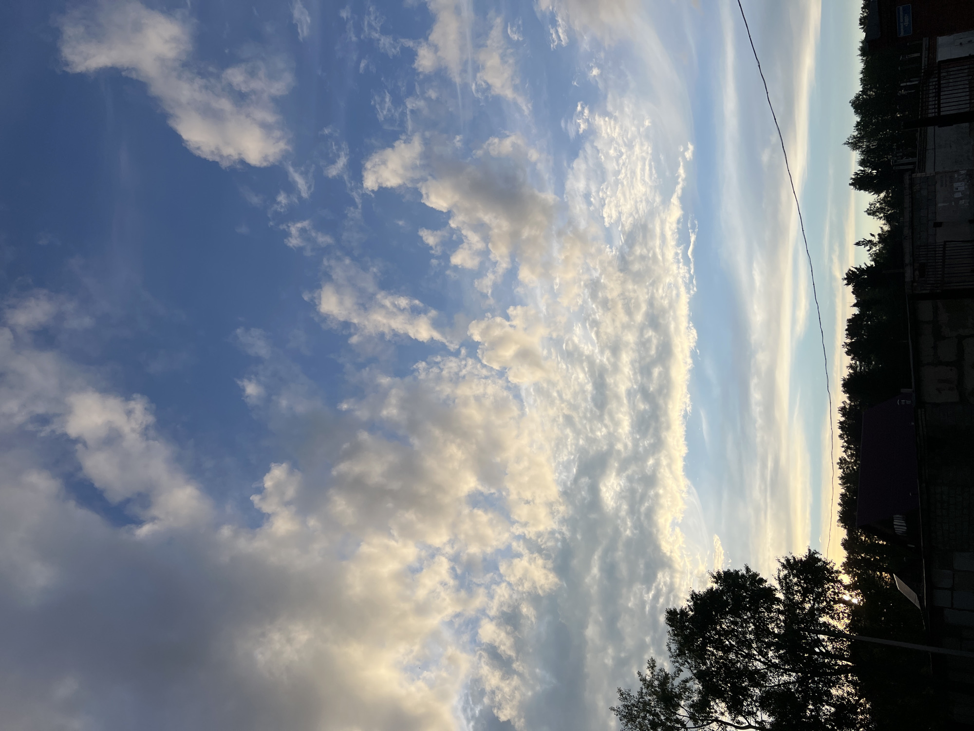 The image is a vertical outdoor shot of a sky filled with clouds. The sky transitions from a deep blue on the left to lighter shades of blue and white streaks on the right. Clouds are scattered throughout, varying in size and density. Some clouds appear puffy and white, while others are more diffused and gray.
The lower portion of the image contains silhouetted outlines of trees and buildings. A power line stretches across the upper right portion of the image.
The location in Chaikovsky, Russia, is inferred from metadata, not visually discernible elements. The time of day appears to be late afternoon or early evening.