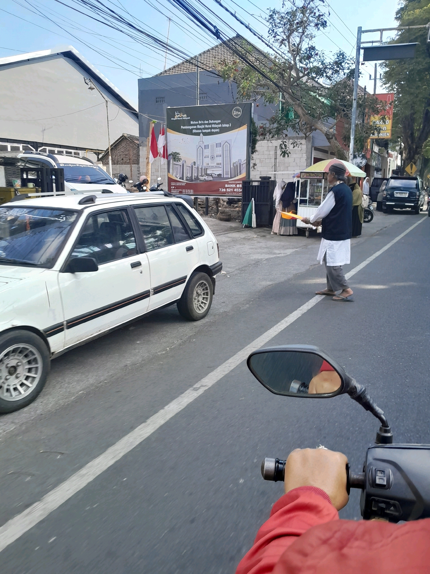 The image shows a street scene in Kepanjen, Indonesia, on a sunny day with a clear blue sky and power lines overhead. In the foreground, the perspective is from a motorcycle, showing a rider's hand in a red jacket gripping the handlebar and a side-view mirror reflecting the rider. To the left, a white hatchback car is parked on the side of the road, partially obscuring the view.
Further down the street, a man in traditional clothing, including a dark vest, white long-sleeved shirt, grey pants, and sandals, walks with a yellow object, possibly a broom or shovel, held in front of him. He appears to be crossing the road. Behind him, a food cart with an umbrella is visible, with people gathered around it, including a woman in a hijab.
A banner advertising the construction of "Masjid Nurul Hidayah" (Nurul Hidayah Mosque) is displayed on a wall. Indonesian flags are also visible. The street is paved and marked with a white line. In the background, buildings and trees line the street, and a dark-colored SUV is seen driving away. The overall impression is of a busy street in a town or city.