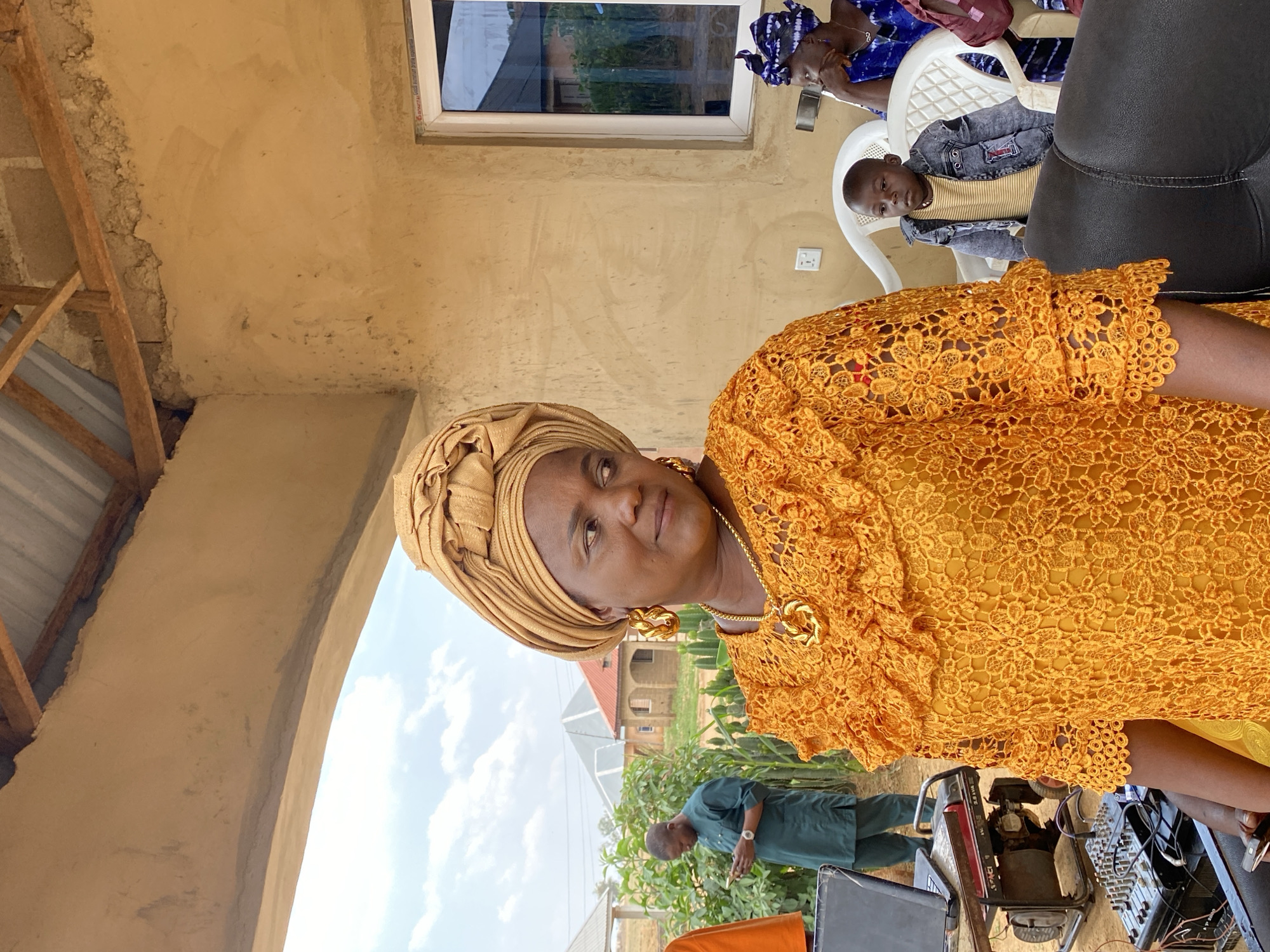 In Barikin Sabon Gidan Kanat, Nigeria, an adult woman wearing a gold-colored lace garment and a matching head wrap is positioned in the foreground. She also sports gold earrings and a necklace. Behind her, a man in a green outfit and another woman in a blue head wrap are visible, seated. A young boy is seated in a plastic chair. To the woman's right is what appears to be audio equipment, and behind her is a view of a building. The image is taken outdoors, under daylight.