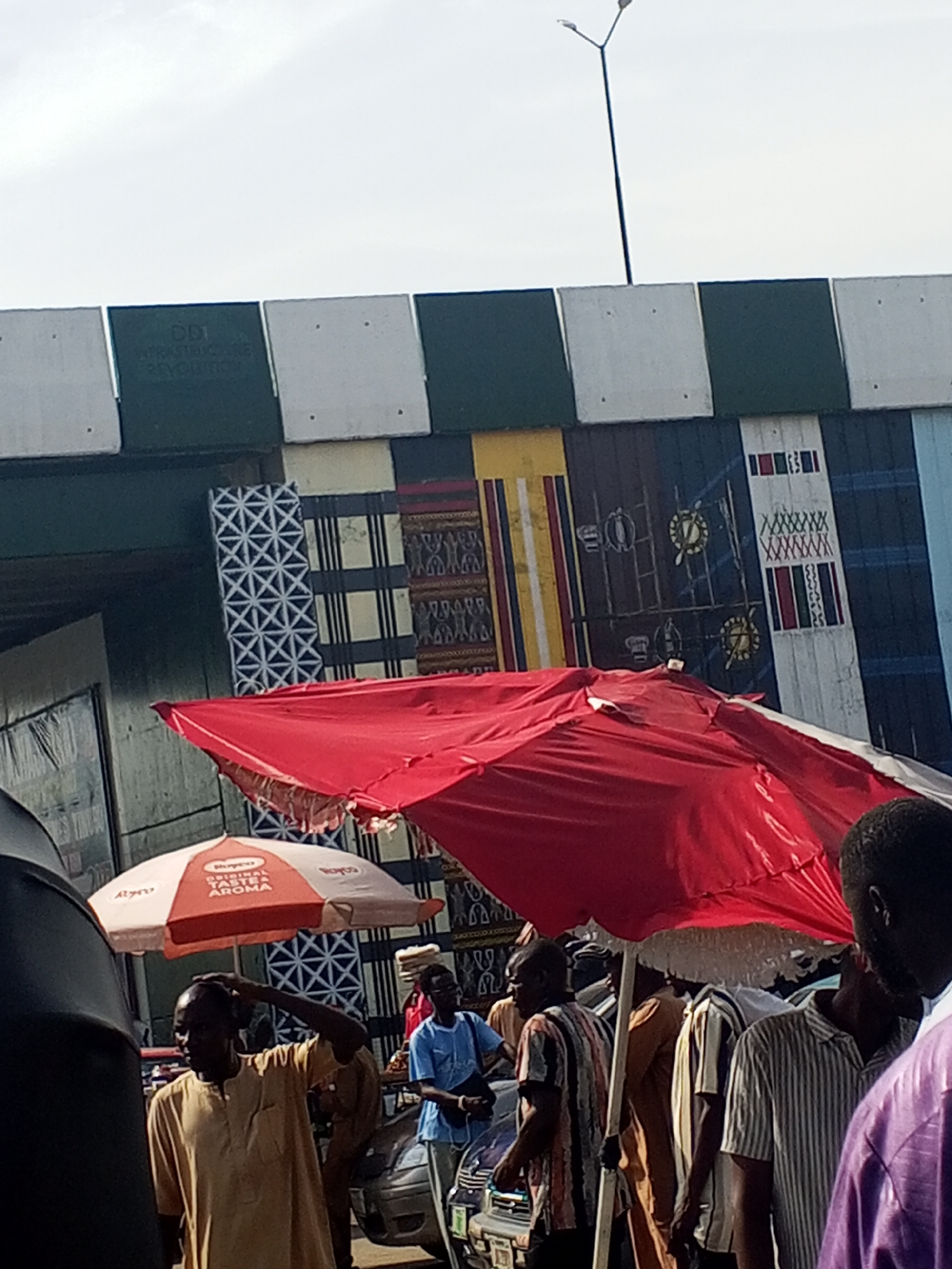 The image depicts a vibrant street scene in Jalingo, Nigeria. The foreground is dominated by a large, bright red umbrella, casting a shadow over a group of people. Beneath the umbrella, a man in a light yellow, long-sleeved tunic is seen scratching his head, his expression unreadable. To his left, another umbrella, this one a mix of orange and white, displays text that appears to be a brand name and possibly a slogan like "original taste aroma."
The background features a colorful mural painted on a wall, with geometric patterns and what seem to be decorative motifs. Above this mural, a concrete structure, likely an overpass or bridge, is visible. The sky is overcast, suggesting a cloudy day, and the lighting indicates it could be daytime. The presence of market stalls, umbrellas, and people suggests a bustling marketplace environment. Several individuals are dressed in various forms of traditional and modern attire. Some are standing, while others are partially obscured by the foreground elements. The overall atmosphere appears to be one of everyday life and commerce in a Nigerian urban setting.