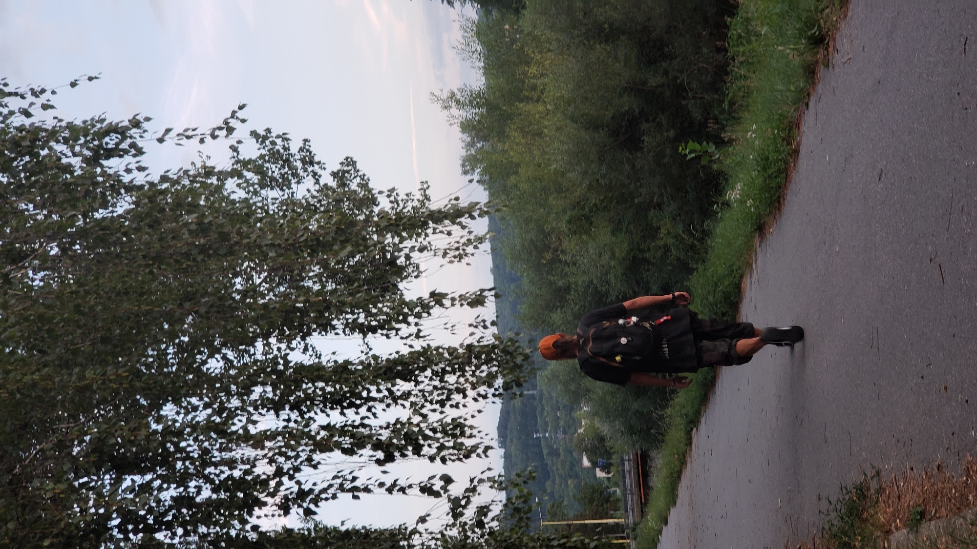 A person wearing a black t-shirt, camouflage shorts, and an orange baseball cap walks away from the camera on a paved pathway. They are carrying a black backpack with several items attached. The pathway is bordered by grass and trees on the right, and on the left, there is a lower grassy embankment with some scattered leaves. In the background, there are lush green trees and hills, and a bridge can be seen on the left side of the frame. The sky is a light blue with some wispy clouds. The image appears to be taken in a park or recreational area within the city of Prešov, Slovakia, possibly near a river or body of water given the vegetation and landscape.