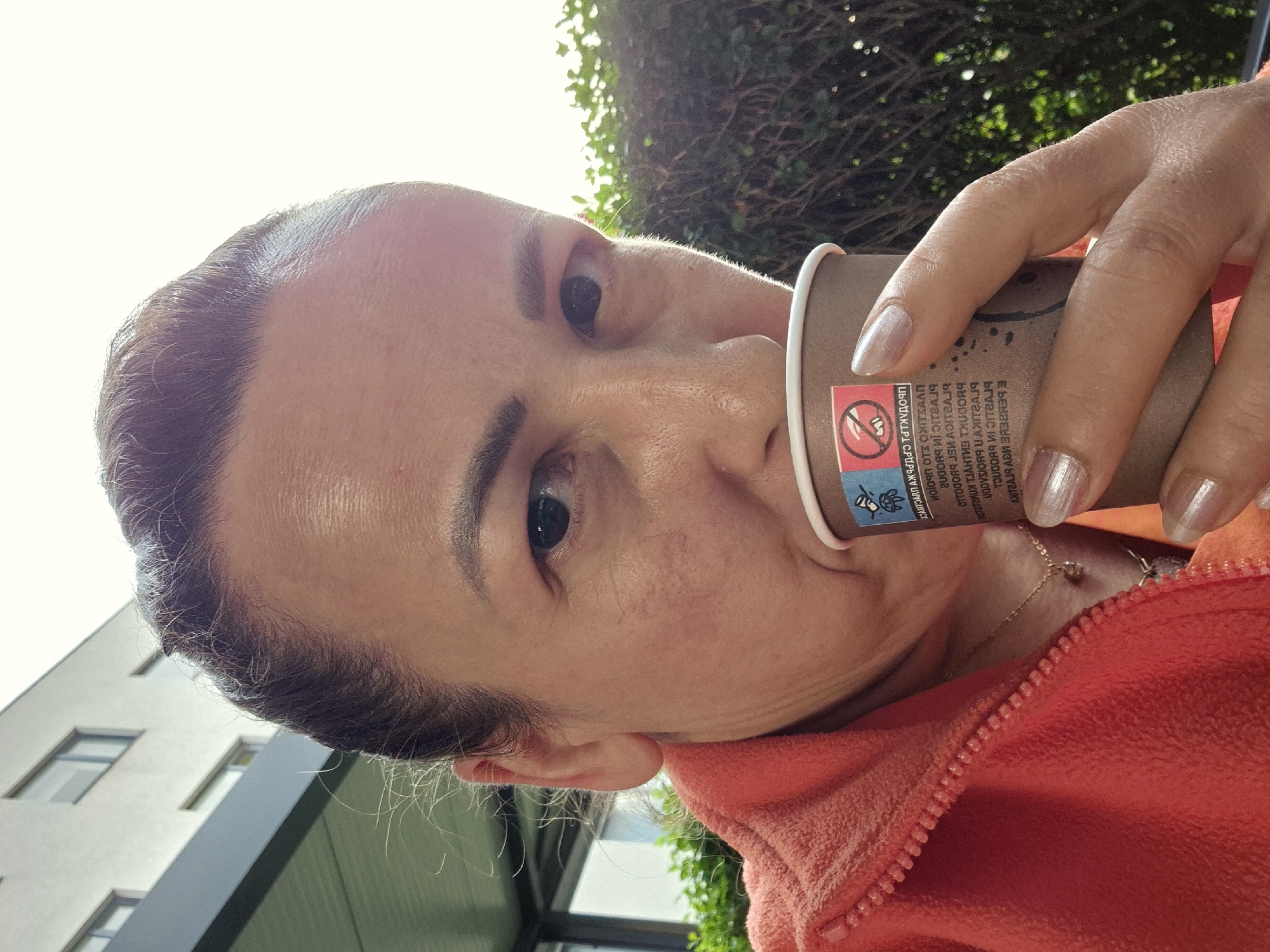 A close-up, selfie-style medium shot captures a woman in an outdoor setting, holding a disposable coffee cup to her lips. She is wearing an orange fleece jacket. Her dark hair is pulled back, and she is looking towards the camera. A small sticker with a prohibition symbol and a turtle is affixed to the coffee cup. In the background, out of focus, is a modern building with windows and some greenery, suggesting an urban environment. The lighting indicates it is daytime, with bright sunlight causing some glare.