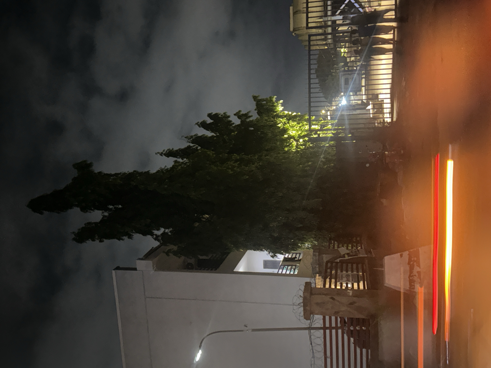 The image depicts a nighttime street scene in Gwarinpa, Nigeria. A tall, slender tree with dense foliage is illuminated by a light source, casting a green glow. To the left, a portion of a modern, light-colored building is visible, featuring a balcony and a street lamp. In the foreground, blurred streaks of red and orange light suggest moving vehicles. A metal gate with vertical bars is prominent in the middle ground, behind which several figures appear to be standing, silhouetted against the background. Further to the right, another section of a building or structure with a rounded top and vertical paneling is visible. The sky is dark and cloudy, typical of nighttime conditions. Barbed wire runs along a low barrier in front of the building on the left.