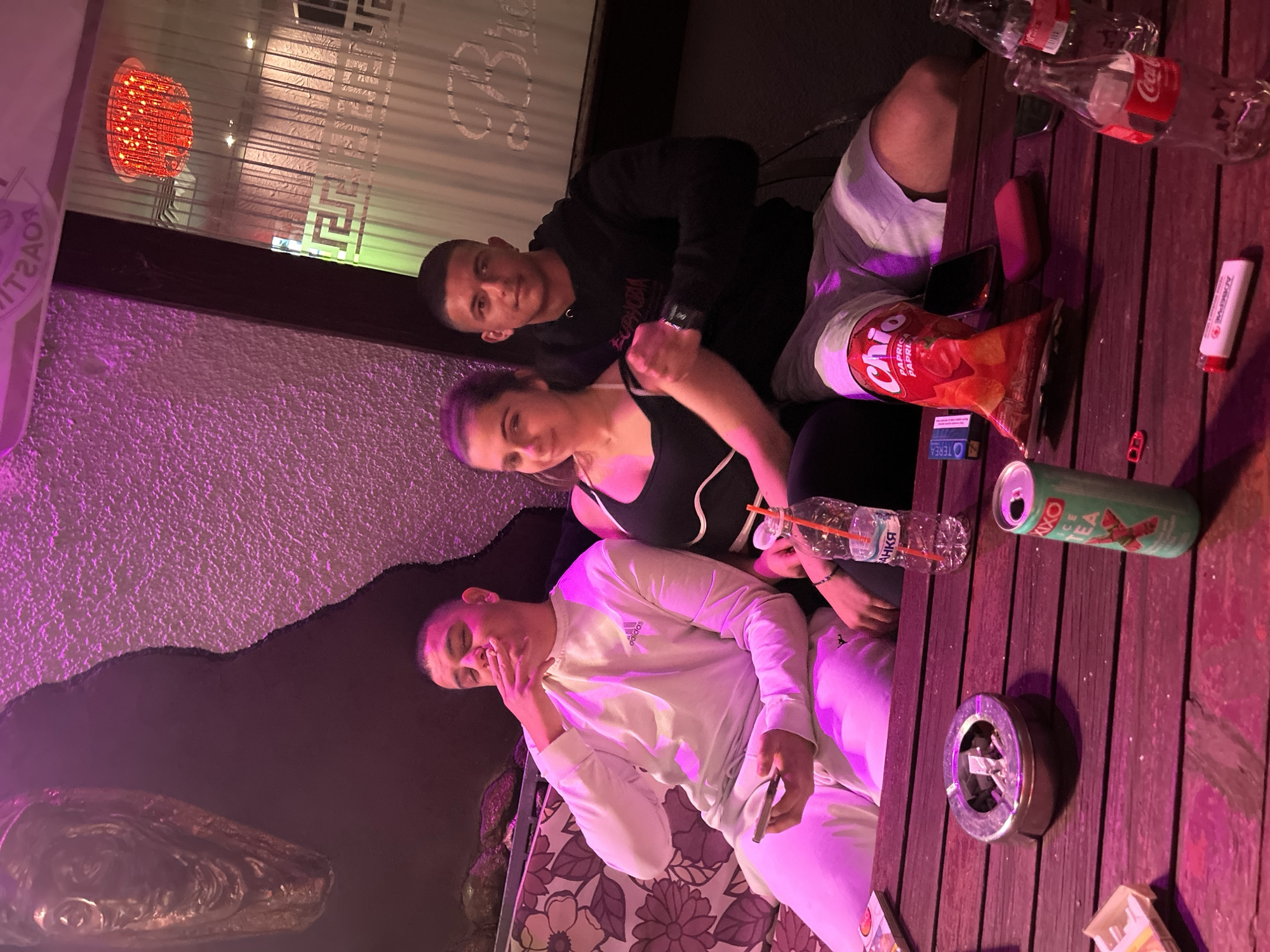 The image shows three young adults gathered around a wooden table in a dimly lit, purple-hued setting, likely an outdoor patio or covered area at night. The lighting casts a strong pinkish-purple glow over the scene.
On the left, a man in a white Adidas tracksuit is partially visible, holding a smartphone and appearing to smoke a cigarette, with his eyes closed or looking downwards. Next to him, a woman wearing a black top looks directly at the camera, with her arm slightly raised. Behind her and to the right, another man, dressed in a black hoodie with "EUPHORIA" visible on the front, also gazes towards the camera.
The wooden table in the foreground is cluttered with items indicative of a social gathering. These include two empty Coca-Cola bottles, a half-eaten bag of Chio Paprika chips, a black smartphone, and a white stick labeled "ACTIVE LOREAL." An open green can of "AXO ICE TEA" is present, alongside an ashtray containing several cigarette butts and a partially visible "OTEREA" cigarette pack. Notably, a clear plastic water bottle with a straw displays the Cyrillic text "БАНЯ," which translates to Bania, providing a specific location cue consistent with Bania, Bulgaria.
In the background, a textured wall is visible, along with a window or glass partition featuring a decorative Greek key pattern and faint, reversed text that reads "ROAST." A red, circular, illuminated lamp adds to the ambient lighting. The overall mood is relaxed and social, suggesting an evening hangout.
