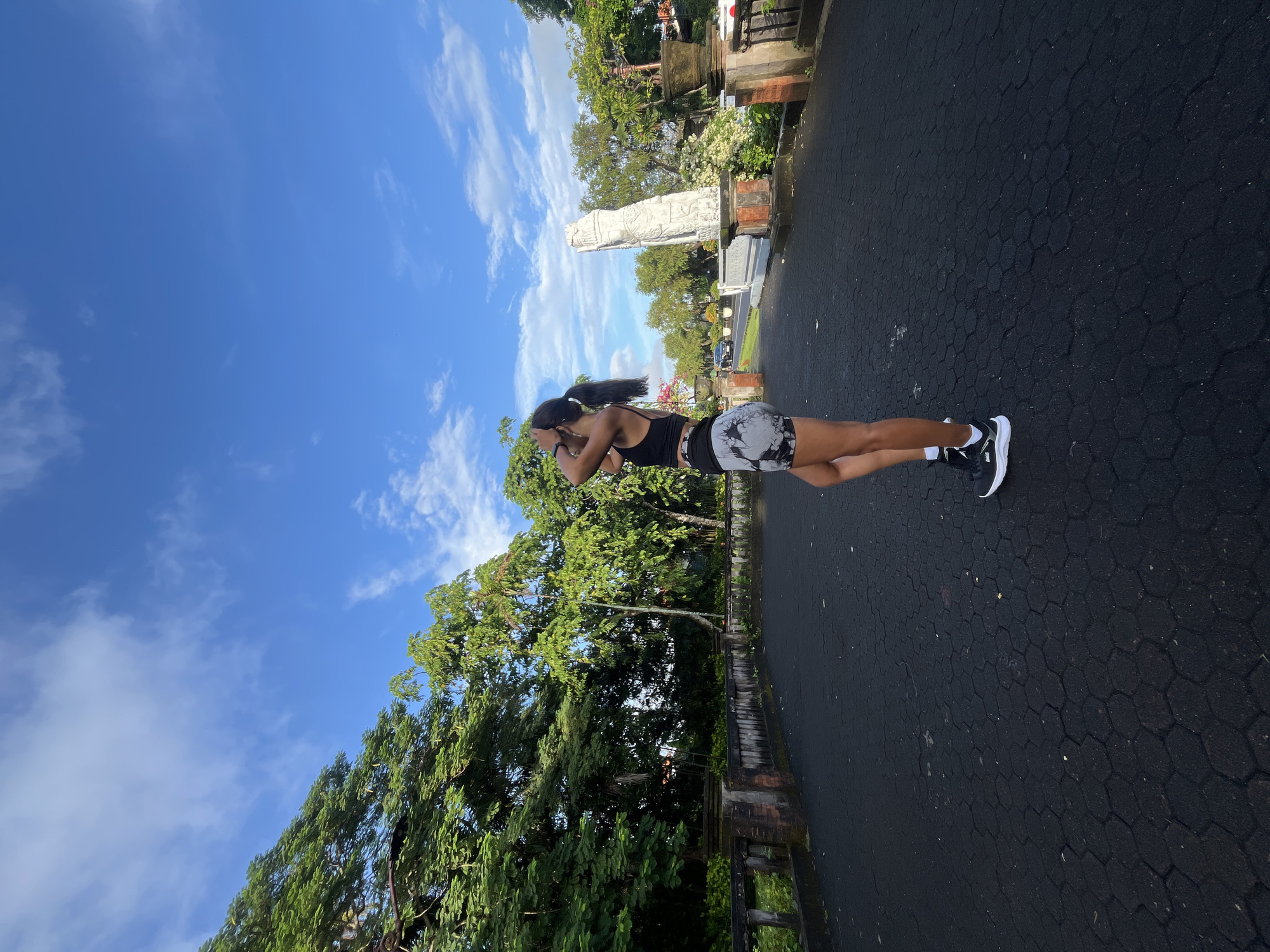 The image, rotated 90 degrees counter-clockwise, depicts a young woman standing outdoors on a paved path. She is seen from the side, facing away from the camera and slightly to the left, with her head tilted upwards. Her dark hair is pulled back into a ponytail. She wears a black sports bra or tank top, black and white patterned athletic shorts, white ankle socks, and black and white athletic shoes. A dark-colored watch or fitness tracker is visible on her left wrist.
The ground she stands on is a dark, possibly asphalt or concrete surface with a distinct hexagonal pattern. To her left and extending into the background are numerous lush green trees and dense foliage, indicating a tropical environment. In the mid-ground, behind the woman, stands a tall, intricately carved white stone statue or monument, featuring traditional Balinese-style elements. A low, dark stone or brick fence or wall runs along the edge of the paved area, separating it from the greenery. The sky above is a vibrant clear blue with scattered wispy white clouds.
The scene is brightly lit by natural sunlight, suggesting either morning or afternoon. The overall setting implies a peaceful outdoor area, possibly a park, temple ground, or pathway in Kuta Selatan, Indonesia. No discernible text is visible in the image.