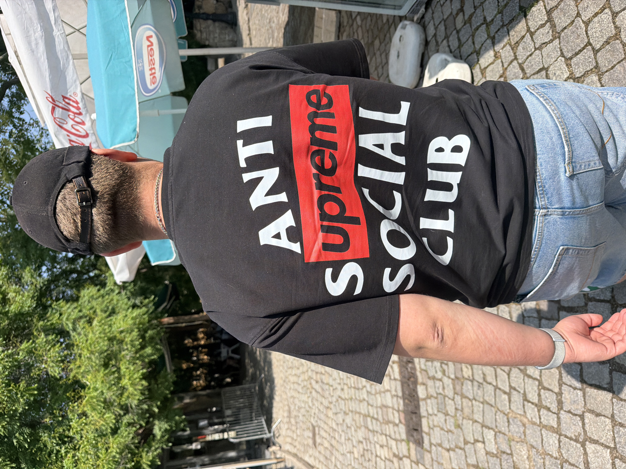 The image, captured in Plovdiv, Bulgaria, depicts a person from behind, standing outdoors on a sunny day. The individual wears a black t-shirt, light blue denim jeans, and a black baseball cap with a brown leather strap and buckle. A silver chain necklace is visible around their neck.
The black t-shirt features a bold design on the back. It reads "ANTI" in large white block letters, followed by the word "Supreme" in white letters set against a prominent red rectangular background, and then "SOCIAL CLUB" also in large white letters. The complete phrase on the t-shirt is "ANTI Supreme SOCIAL CLUB". The person's right arm is visible, wearing a light-colored wristband, likely from a smartwatch.
The setting is an outdoor public area, with the ground paved in cobblestones. In the background, green trees are visible under a clear, bright sky, casting distinct shadows that suggest strong daylight. Two large outdoor umbrellas are also present: one white, partially displaying a red "Coca-Cola" logo, and another light blue umbrella featuring a mirrored "Nestle" logo. These details indicate the location is likely an outdoor cafe or a commercial area within the city. The overall scene suggests a casual, daytime atmosphere.