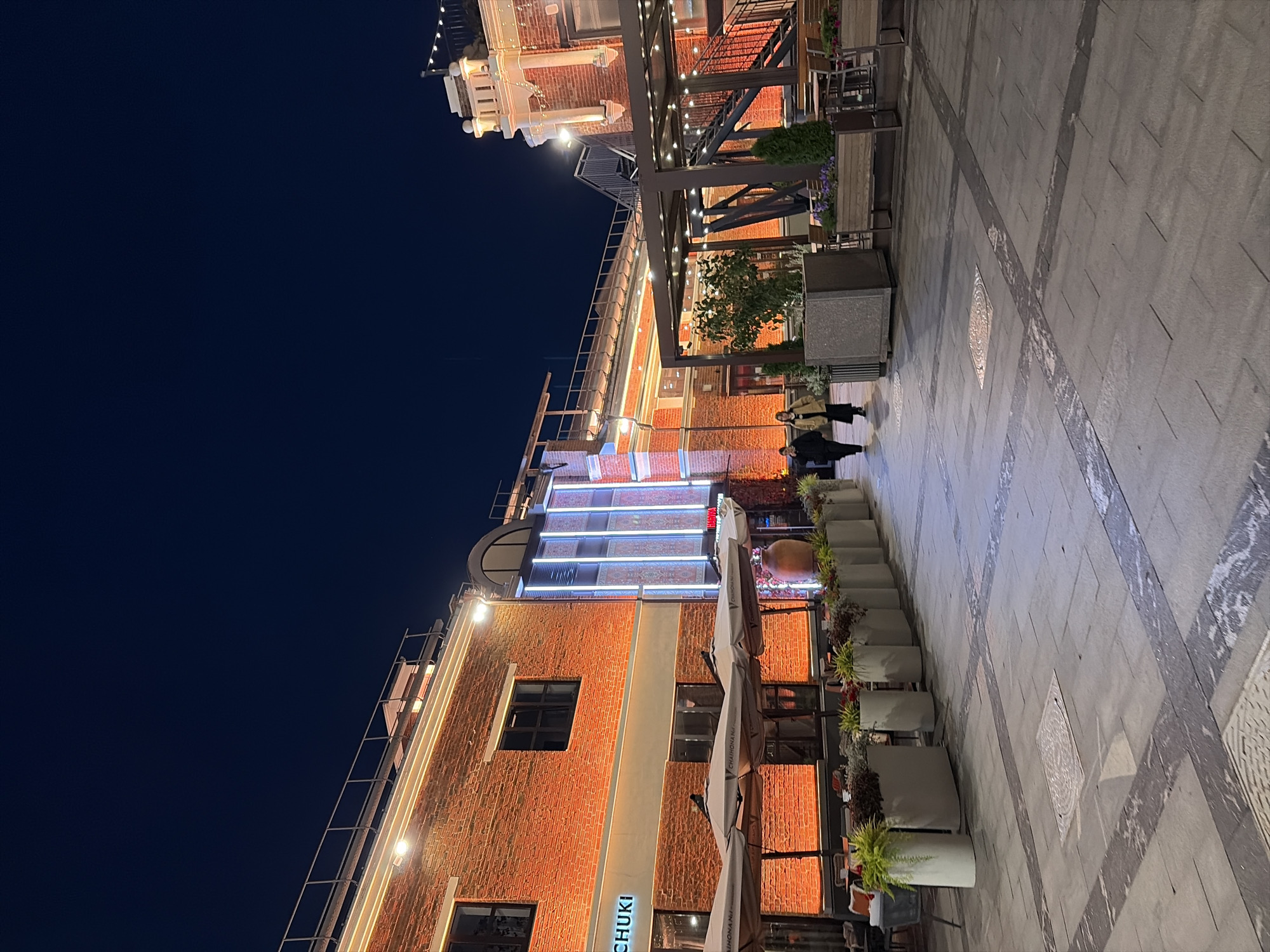 This is a nighttime exterior shot of a brick building with outdoor seating and a courtyard. The building has warm orange lighting and is illuminated with neon signs and string lights. There are several tables and chairs under umbrellas, suggesting an outdoor dining area. Planters with greenery line the walkway. Two individuals are walking away from the camera in the distance. The ground is paved with large stone tiles. The architecture of the building includes decorative elements and an external staircase. This scene is consistent with an outdoor area of a restaurant or entertainment venue in Moscow, Russia.