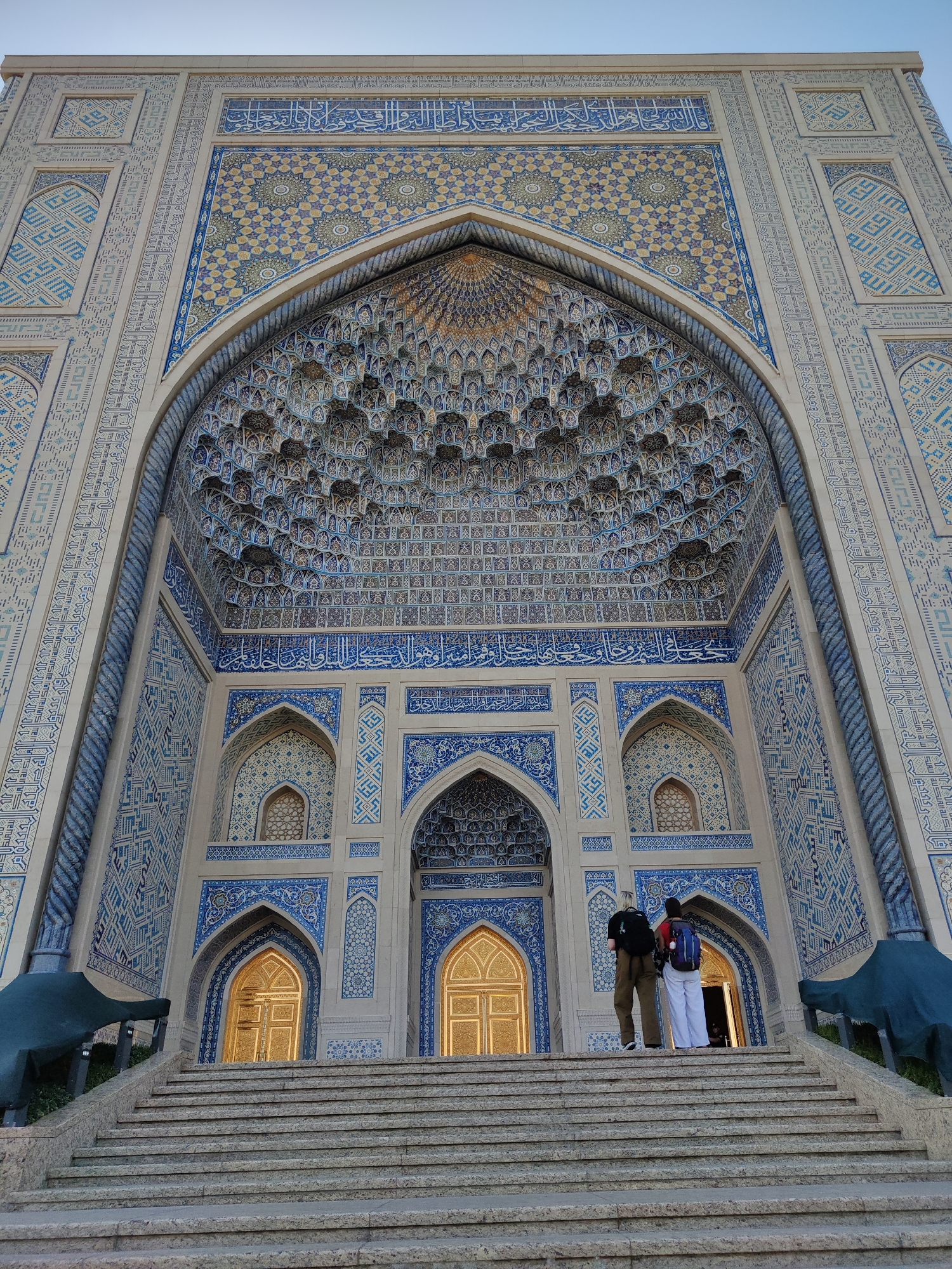 The image captures the entrance to the Khast Imam complex in Tashkent, Uzbekistan. The architecture features intricate Islamic geometric patterns and Arabic calligraphy in shades of blue and white against a light-colored stone facade. A grand, arched portal dominates the center, adorned with detailed muqarnas work. Two individuals, a man and a woman, stand on a wide stone staircase leading up to the entrance. Both are carrying backpacks, suggesting they are tourists. The man is wearing khaki pants and a dark shirt, while the woman is in white pants and a red shirt. Golden doors are visible through the arched entryway. The sky above is clear and pale. The overall scene conveys a sense of grandeur and historical significance.