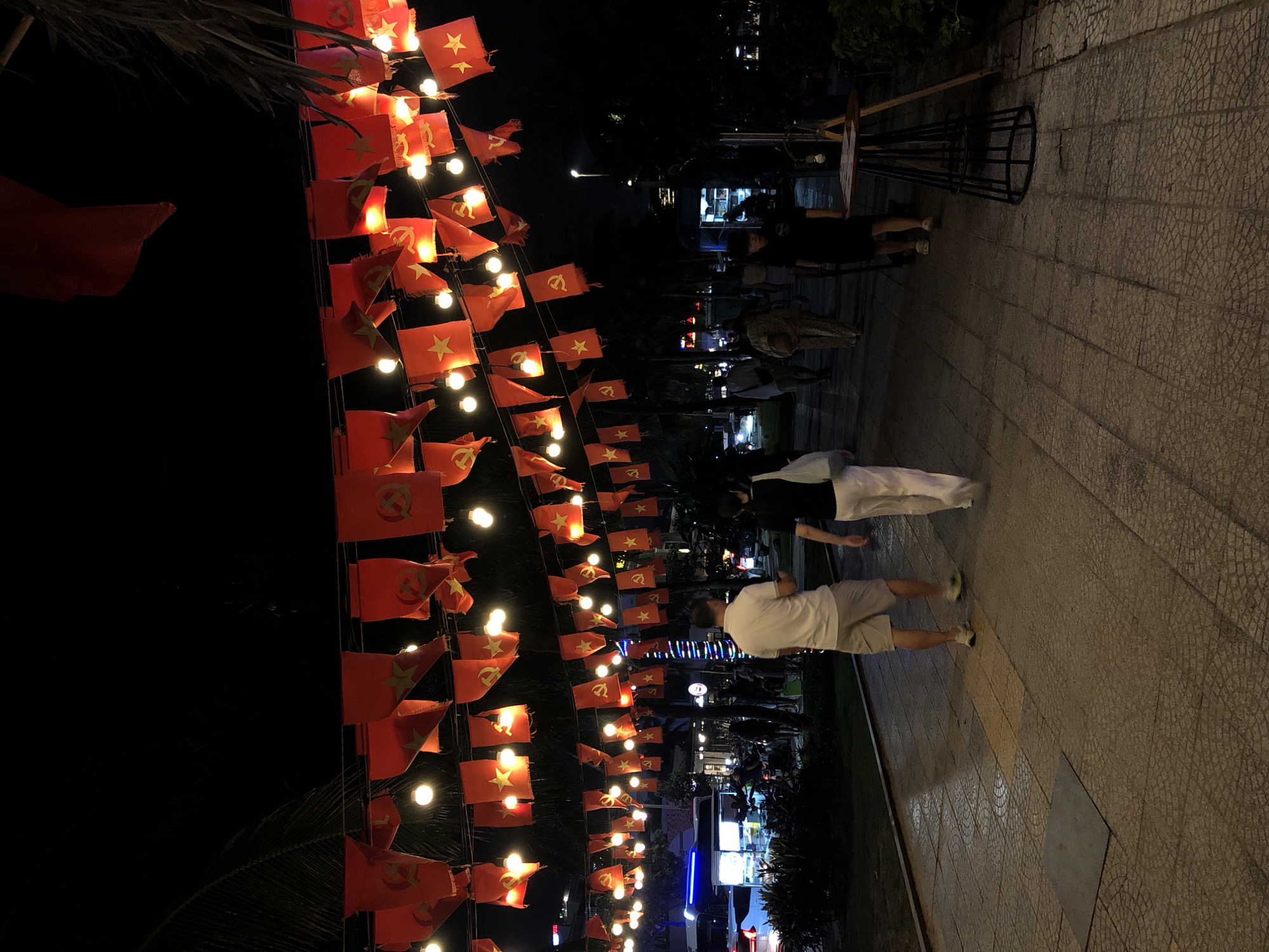 The image captures a street scene at night in Da Nang, Vietnam. Red flags with yellow stars and hammers and sickles adorn strings stretched across the street, interspersed with illuminated light bulbs, creating a festive atmosphere. Several people are walking along a paved walkway. In the foreground, two individuals are seen from the back, one wearing a white polo shirt and shorts, and the other in black attire. Further down the walkway, more people are visible, along with parked vehicles and buildings in the background. The lighting suggests it is after sunset.