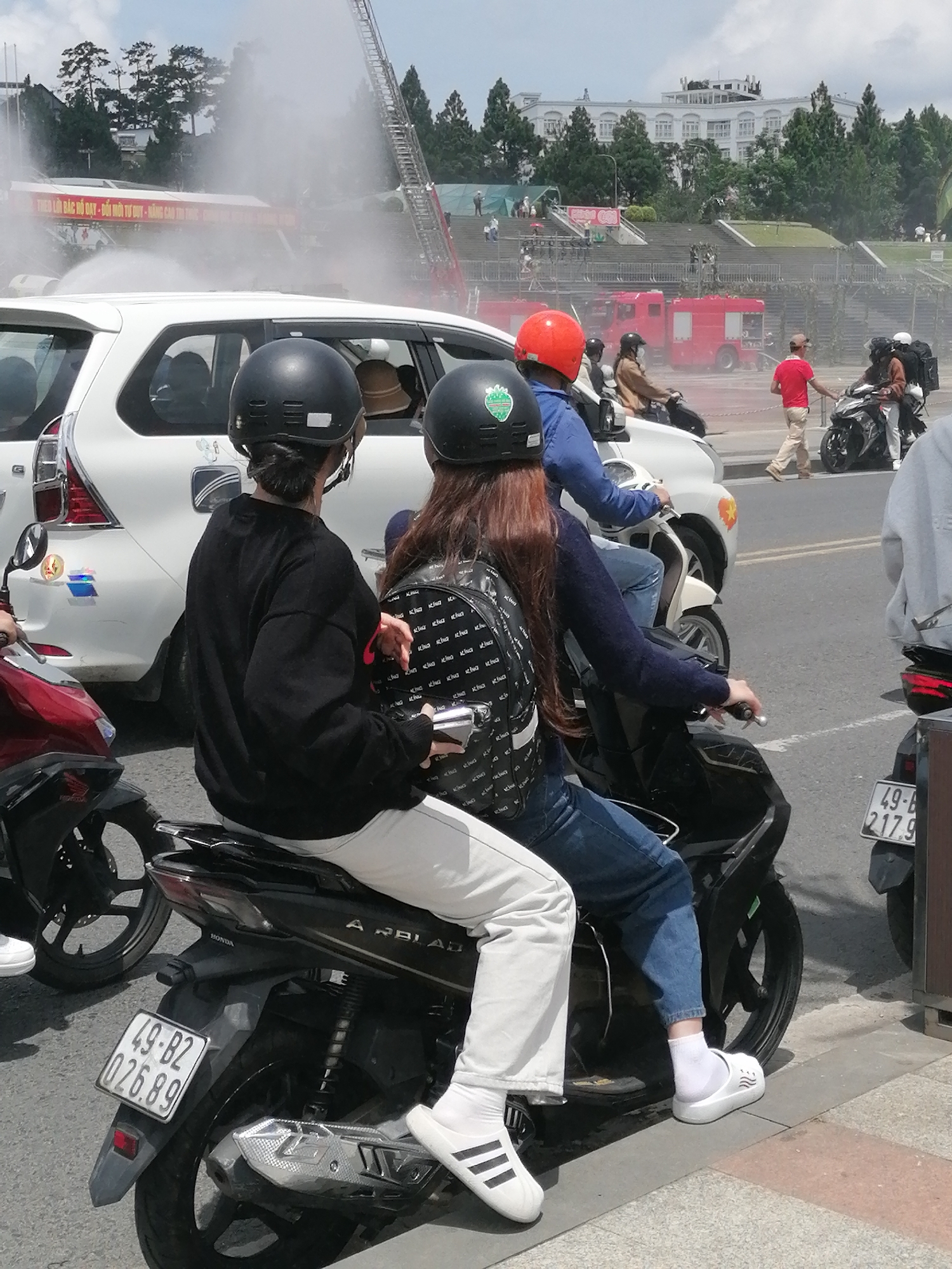The image depicts a street scene in Da Lat, Vietnam, possibly during a public event or demonstration. In the foreground, two individuals wearing helmets are seated on a dark-colored scooter, the passenger on the left wearing a black long-sleeved shirt and white pants, and the passenger on the right wearing a blue jacket and blue jeans. Both are looking forward. Behind them, a white SUV is parked, and further back, red fire trucks are visible, with water spraying from a fire hose onto a building or structure. Other vehicles, including motorbikes and a car, are present on the street. In the background, there is a large white building with multiple windows, and steps leading up to it, surrounded by green trees. The sky is cloudy. The license plate on the scooter is clearly visible and reads "49-B2 026.89". The overall atmosphere suggests an active event taking place in a public area of the city.