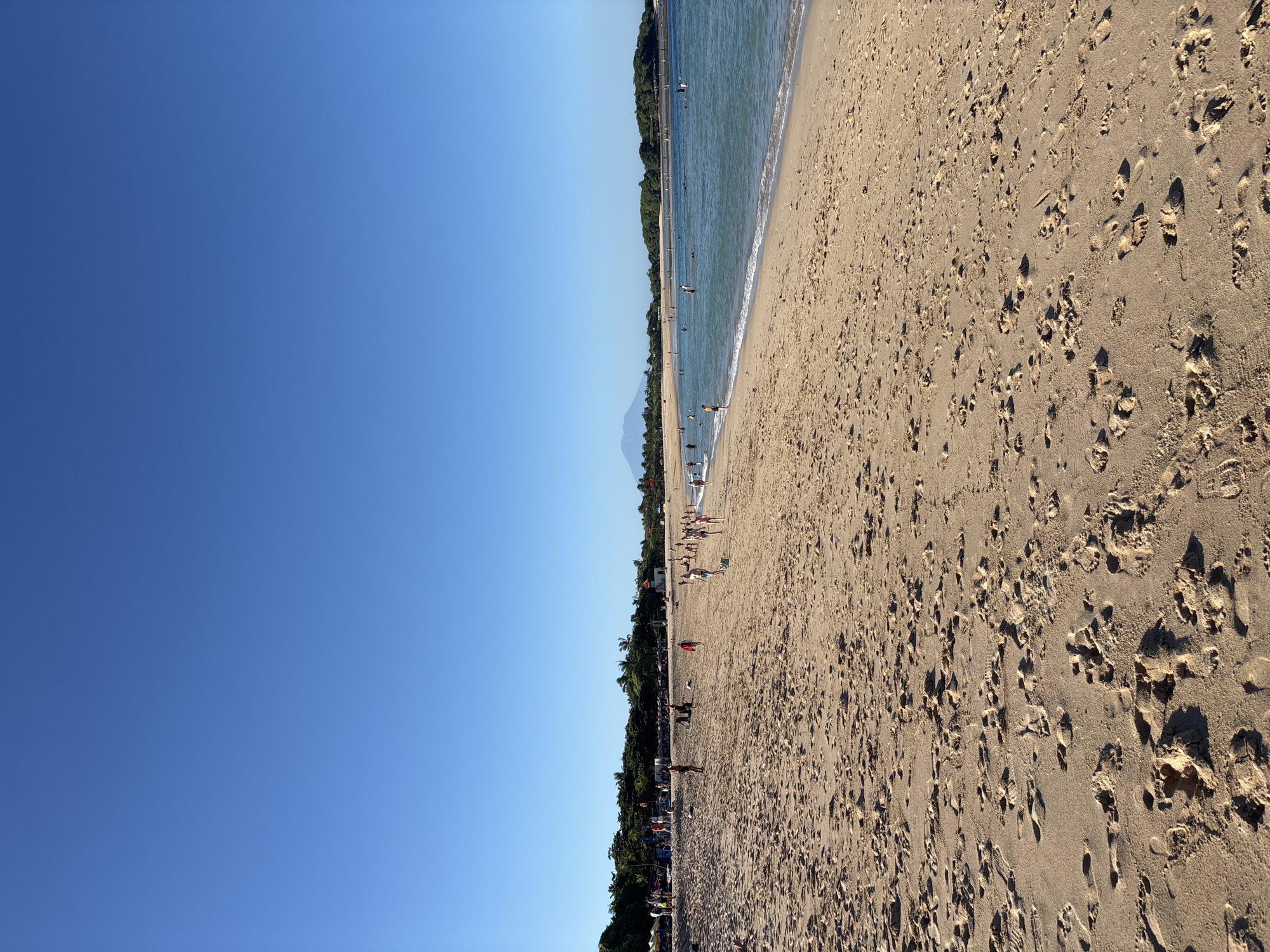 The image captures a beach scene in Kuta Selatan, Indonesia. A clear blue sky fills the left portion of the frame. The beach occupies the right side, showing a tan-colored sandy expanse covered with numerous footprints. The shoreline is visible in the middle ground, where gentle waves meet the sand. People are dispersed along the beach and in the water, engaging in activities such as swimming and walking. A line of trees and vegetation borders the beach, providing a backdrop to the scene. A distant mountain is observed on the horizon.