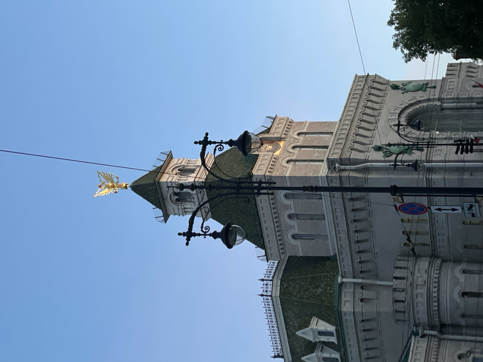 This image captures a close-up view of the Singer House (Dom Knigi) in Saint Petersburg, Russia. The iconic building, situated on Nevsky Prospekt, is characterized by its grand architecture. Prominently featured is the glass dome topped with a golden double-headed eagle, a symbol of Russia. The building's facade is constructed from light-colored brick, with decorative elements and arched windows. A distinctive green tiled roof is visible, adding to the building's unique aesthetic. In the foreground, a black lamppost with ornate detailing and a glass lantern stands silhouetted against the clear blue sky. Several traffic signs, including a red and blue circular sign with a diagonal red line, are also present near the lamppost. The image is taken from a low angle, emphasizing the height and grandeur of the Singer House.