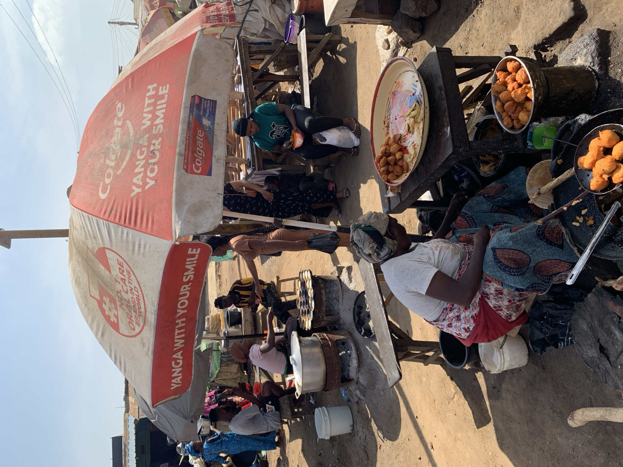Here is a description of the image:
The image shows a busy street scene in what is described to be Rafin Par, Nigeria. Several people are visible, with a group clustered around what appears to be a food preparation and sales area. One woman wearing a white shirt and patterned skirt is seated near a cooking setup with pots and pans. Another woman is seated nearby under a white and red umbrella, and several other individuals are milling around the area. There are fried dough-like foods in containers and pans. The setting appears to be an outdoor market or street food area. A Colgate-branded umbrella is present in the scene. There is no clear indication of criminal behavior or illegal activity.