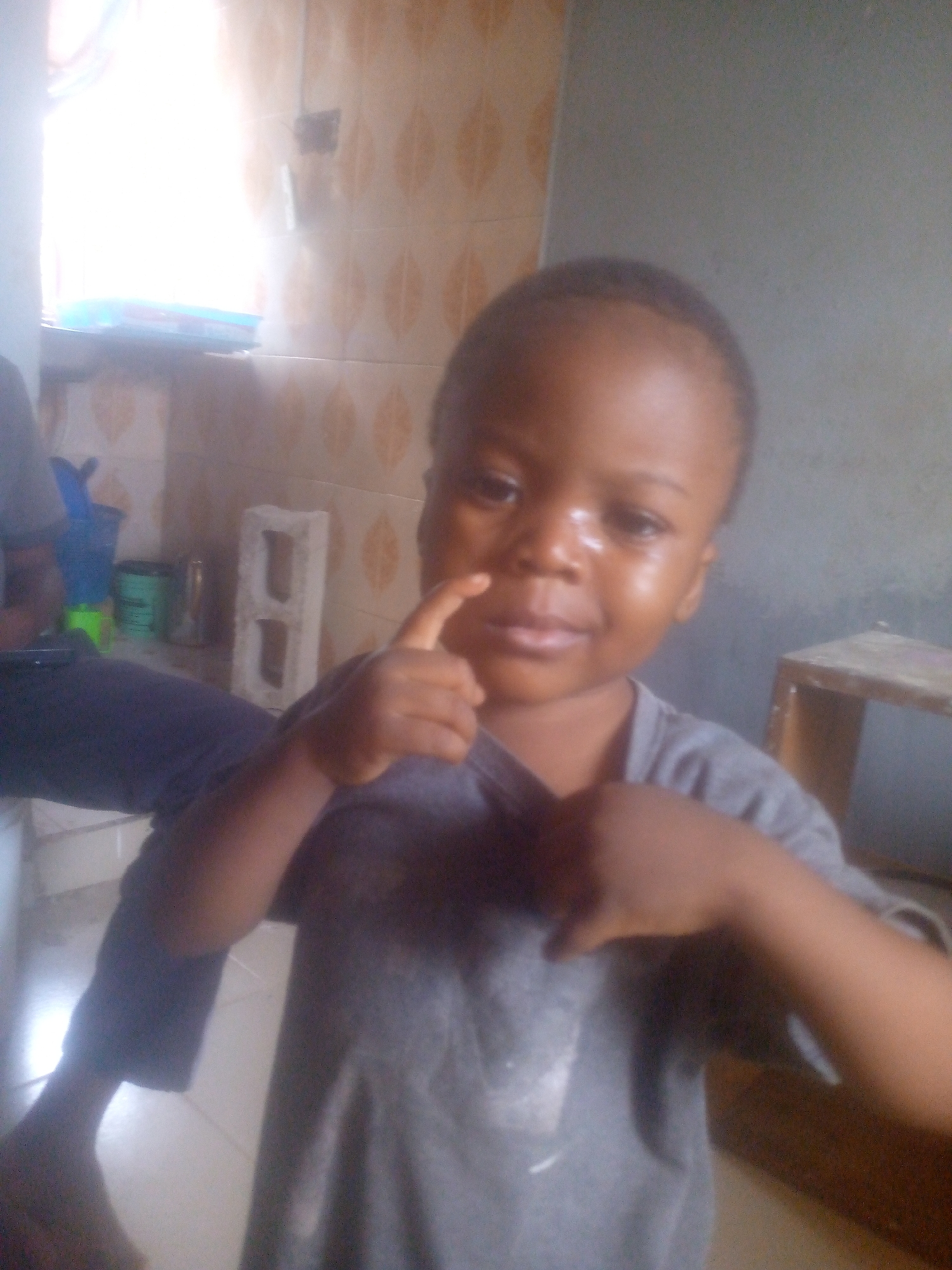 A close-up, slightly blurry indoor photograph captures a young child, likely a toddler, as the central subject. The child, with dark skin and short hair, faces the camera directly, wearing a plain grey t-shirt. Their right index finger is raised and touching their nose or upper lip, while their left hand rests on their chest, gently gripping their shirt. A subtle sheen is visible on the child's face, possibly from light reflection or sweat.
To the left, a portion of another person's leg, clad in dark blue trousers, and a lower arm are visible. The background is an indoor space, featuring light brown patterned tiles on one wall and a plain greyish-green wall on the other side. A bright, overexposed window on the upper left casts strong daylight into the room, obscuring items on its sill. Below the window, on a surface, a blue plastic cup, a green container, and a white, hollow rectangular block are present. On the right side of the image, part of a wooden stool or small table can be seen. The overall impression is a casual, daytime moment. No visible text is present in the image.