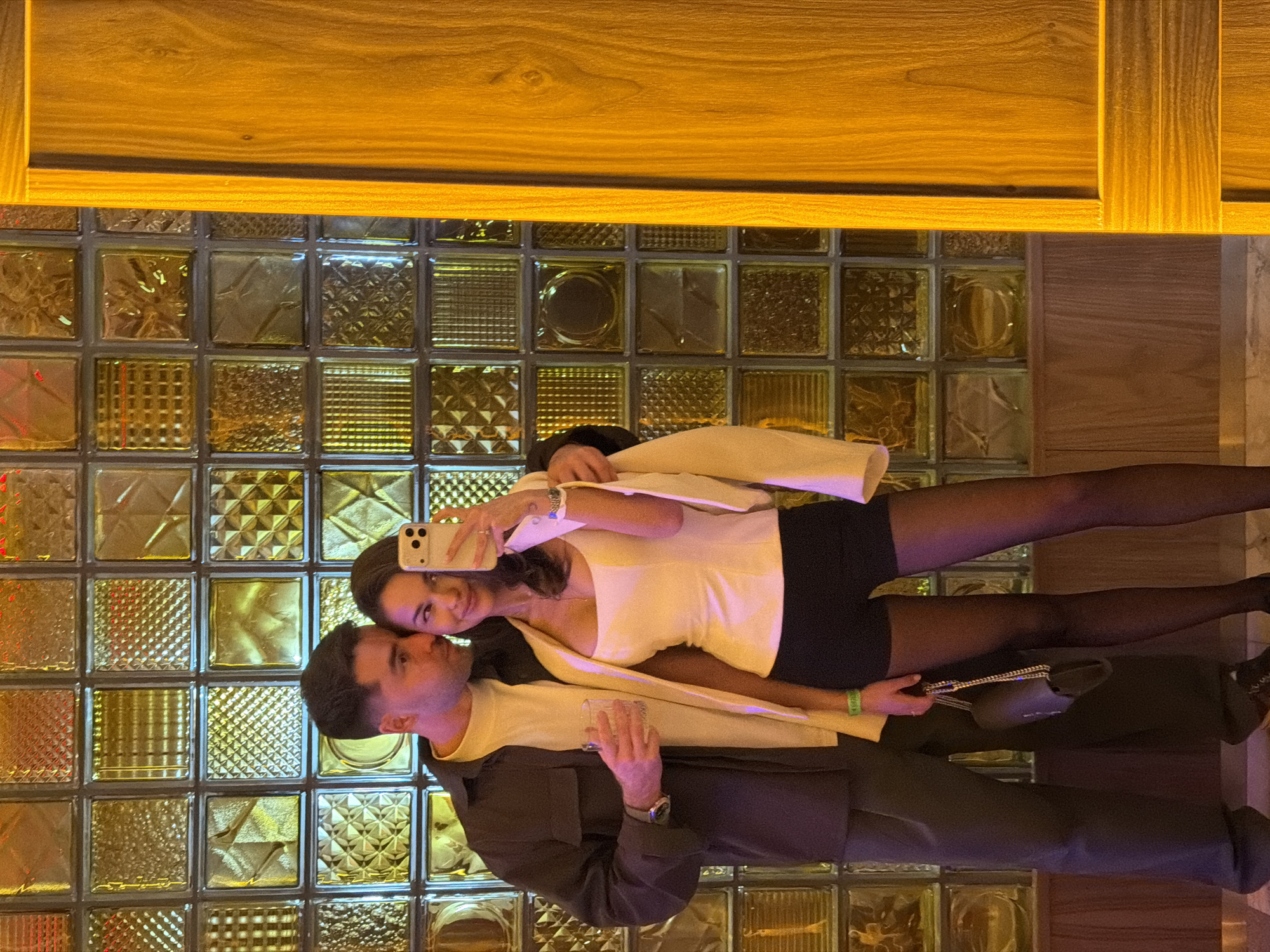An image depicts a couple taking a selfie in a stylish indoor setting in Krasnoyarsk, Russia. The woman, positioned on the right, is holding a smartphone (likely an iPhone) up to capture the photo, looking towards the camera with a gentle smile. She is dressed in a white sleeveless top, a black mini-skirt, and sheer black tights, with a white blazer casually draped over her shoulders. Rings adorn her fingers, and a watch is visible on her left wrist, along with a light green wristband on her right. A black handbag with a silver chain strap rests near her.
The man stands to her left, gazing towards the camera or the woman, also with a subtle smile. He wears a light yellow or cream t-shirt under a dark brown jacket and dark trousers. He holds a clear glass with a beverage in his left hand, and a watch is visible on his wrist.
The background features a distinctive wall constructed from numerous square glass blocks, each exhibiting different embossed patterns and textures. These blocks are illuminated from behind, creating a warm, golden-amber glow that bathes the scene, suggesting an evening or nighttime ambiance. A smooth wooden panel or beam extends across the top of the frame, also reflecting the warm light. The overall mood appears intimate and relaxed, indicating a pleasant social outing. There is no discernible text on clothing or visible objects besides the phone's branding.