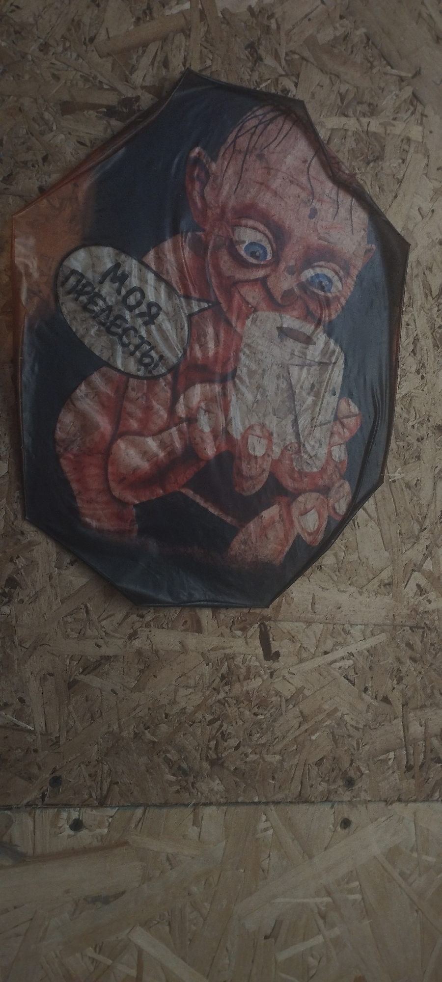 The image shows a printed umbrella hanging on a wooden wall. The umbrella features a stylized drawing of the character Gollum from "The Lord of the Rings," with blue eyes and a speech bubble containing the Russian text "Моя прелесть!" which translates to "My precious!". The wall appears to be made of oriented strand board (OSB) and is likely located indoors, as indicated by the flat, consistent lighting. The specific location in Покосное, Russia cannot be determined from the image alone.