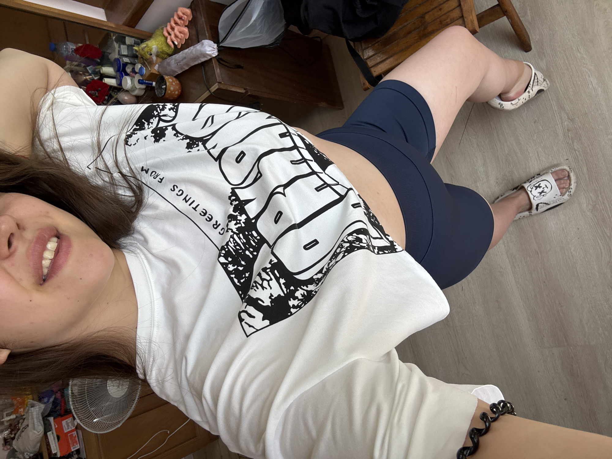 The image shows a person lying down, with their head tilted up towards the camera. They are wearing a white t-shirt with a black graphic print that includes the text "GREETINGS FROM". They are also wearing dark blue biker shorts and patterned slides. The background includes a wooden shelf with various items, a fan, and a wooden chair. The floor appears to be made of laminate. The lighting suggests it is daytime. The person's expression appears to be a smile, and the overall mood of the image seems casual and relaxed.