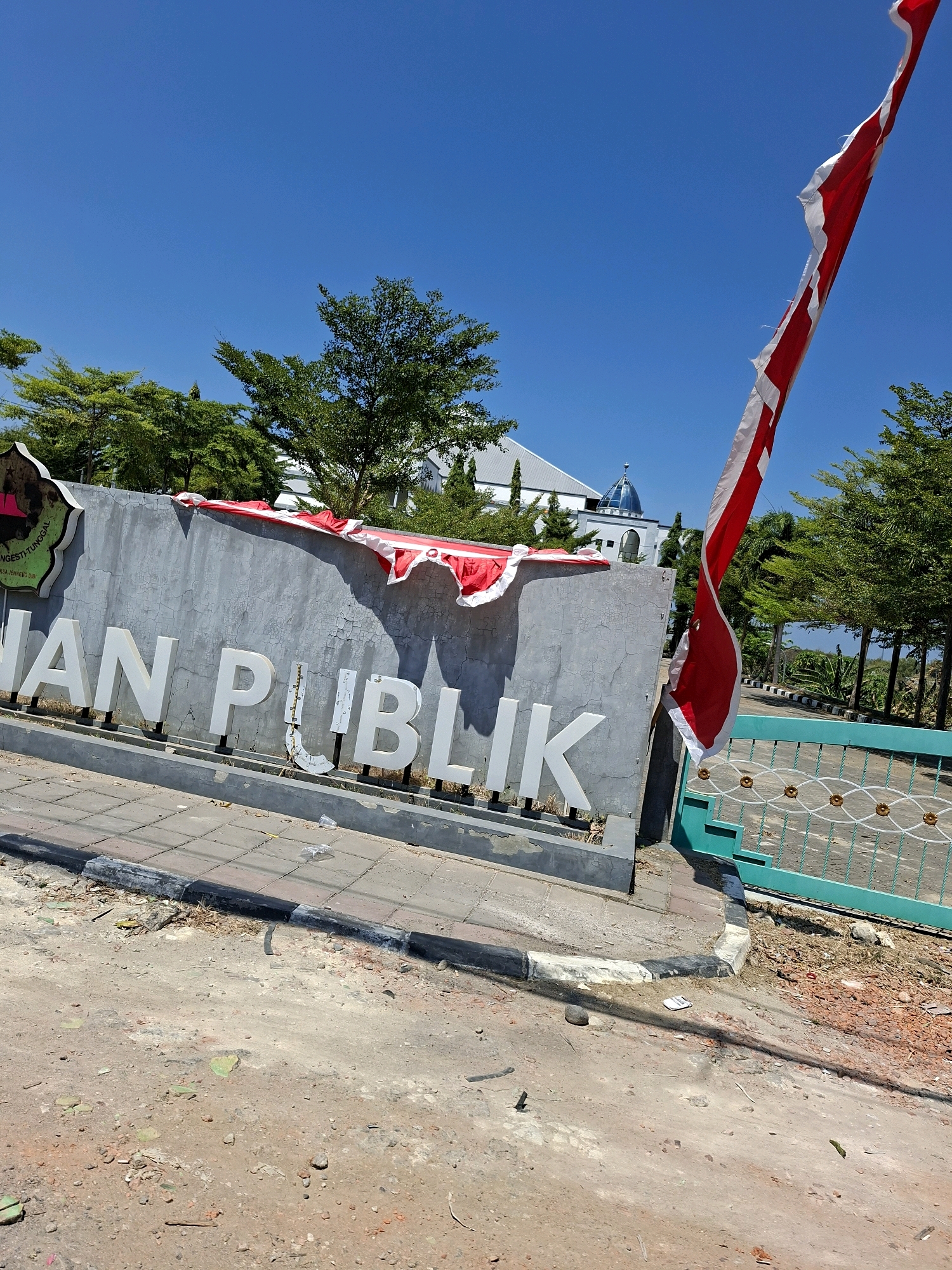 The image shows a concrete wall with large white lettering that spells "AN PUBLIK." A red and white flag, torn and frayed, is draped over the top of the wall and is also attached to a flagpole on the right side of the image, extending upwards into a clear blue sky. The wall is situated next to a sidewalk made of concrete tiles, bordered by a curb. The ground in front of the wall is rough and appears to be under construction, with loose dirt, small rocks, and scattered debris. To the right of the wall, a turquoise-colored metal gate with a decorative pattern is visible. In the background, there are several trees, a building with a blue dome and white structures that resemble a mosque or a public building, and a clear blue sky. The overall scene suggests an outdoor public space, possibly an entrance to a facility, in Ceguk, Indonesia.
