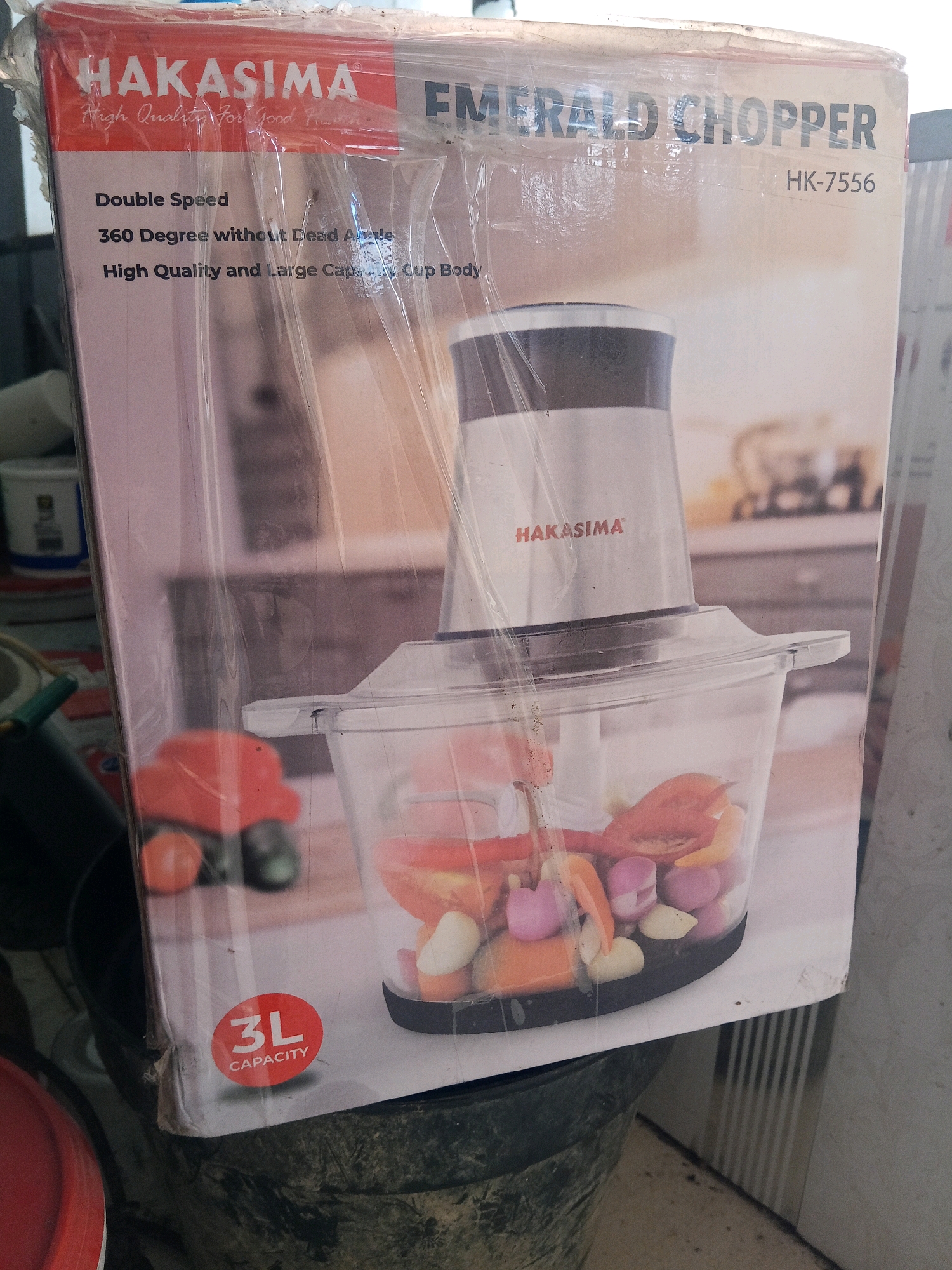 An outdoor scene in Jinang Kampung Pansur, Indonesia, features a partially visible product box for a "HAKASIMA EMERALD CHOPPER HK-7556" positioned in the foreground. The box is wrapped in plastic and shows images of a food chopper with ingredients like onions and tomatoes inside its container. Text on the box indicates features such as "Double Speed," "360 Degree without Dead Angle," and "High Quality and Large Capacity Cup Body." A circular "3L CAPACITY" logo is also visible. The background is out of focus but suggests an urban or semi-urban environment with buildings and possibly other objects, one of which appears to be a white bucket with a blue label. The overall lighting is natural, indicating daylight. There are no people clearly visible in the frame.