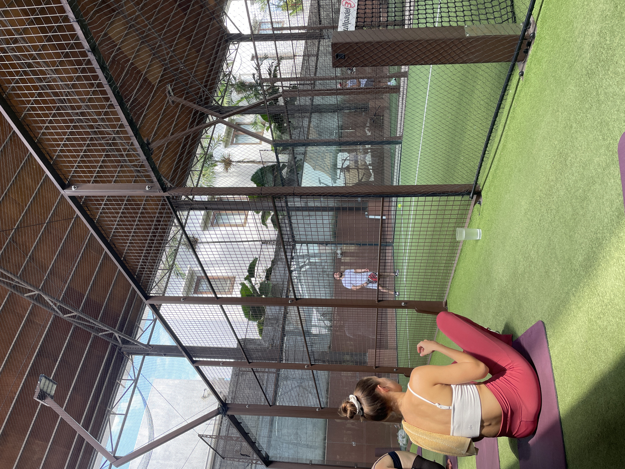 An overhead shot captures a padel tennis court in North Kuta, Indonesia. The court features a green artificial turf surface, enclosed by glass panels and metal mesh fencing, with a sloped, mesh-covered roof overhead. Two men are actively playing padel, one in the foreground on the left and another further back on the right. In the foreground, a woman sits on a purple yoga mat, her back to the camera, wearing a white sports bra and pink leggings, with a yellow towel draped over her shoulder. Another woman, partially visible on the far left, is also seated on a yoga mat. The scene is bathed in sunlight, casting shadows on the court.