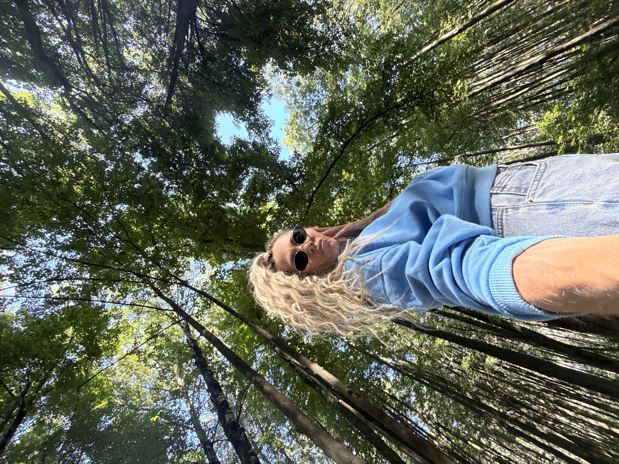 The image depicts a low-angle shot of a young woman with blonde, curly hair tied in a high ponytail. She is wearing sunglasses, a blue sweatshirt, and light-wash denim jeans. The woman is standing in a forest with tall trees that create a canopy overhead, with patches of blue sky visible through the leaves. Her left hand is touching her face. The lighting suggests it is daytime. The location is specified as Dumino, Russia.