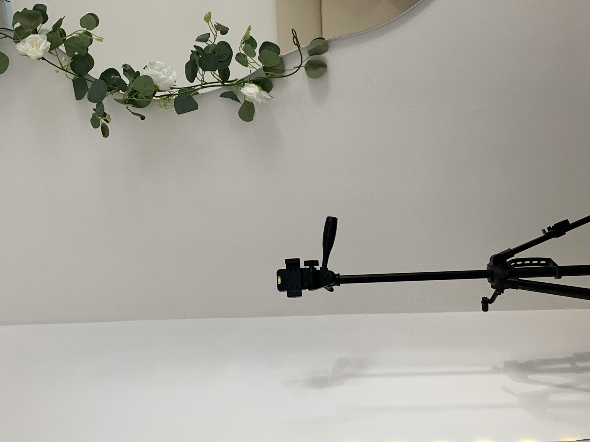 The image shows a tripod in a room with white walls. A decorative garland with white flowers and green leaves is attached to the wall near the top left corner. A portion of a round object, possibly a mirror, is visible behind the garland. The tripod is black and occupies the central and right areas of the image. The foreground is a white surface. The location is unknown, and there is no information to suggest that the image was taken in Фоки, Russia.