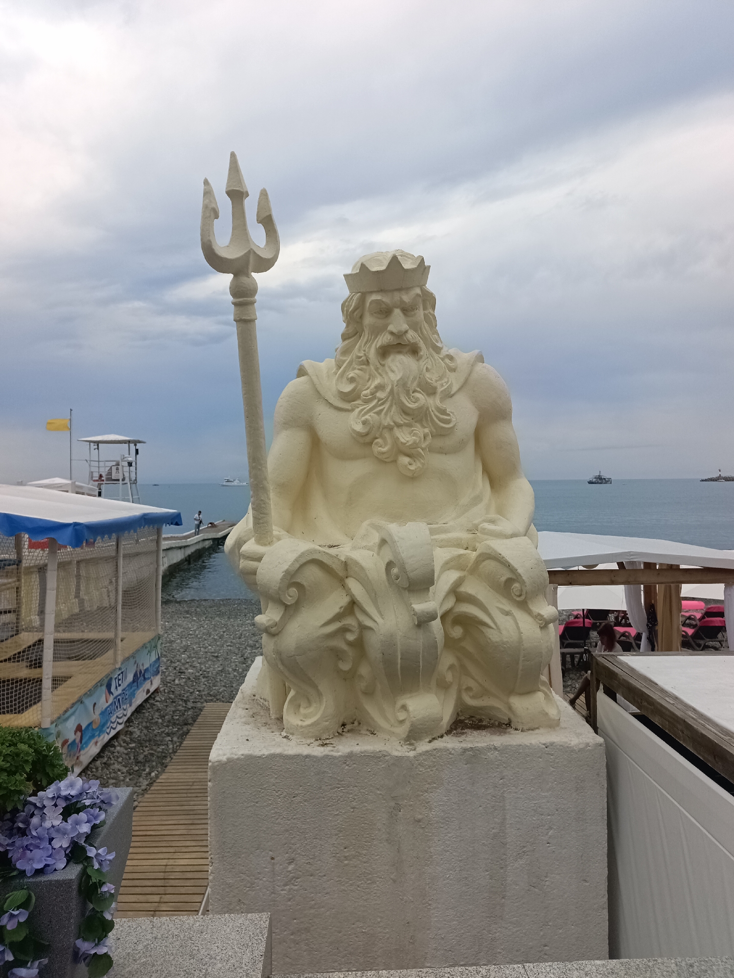 This is a medium shot of a stone statue of a seated man with a crown and beard, holding a trident. The statue is light-colored and appears to be made of concrete or a similar material. It is positioned on a stone pedestal. In the background, there is a beach with a body of water, likely the Black Sea, given the location of Sochi, Russia.
To the left of the statue, there is a beach area with a wooden walkway, a small structure with a blue and white canopy, and what looks like a children's play area. Further back, a lifeguard tower with a yellow flag is visible on a pier extending into the water. There are a few people on the pier and in the distance.
To the right of the statue, there are white canopies providing shade over rows of empty lounge chairs, some with pink cushions. The sky is overcast with gray clouds. The overall scene suggests a public beach or resort area in Sochi.