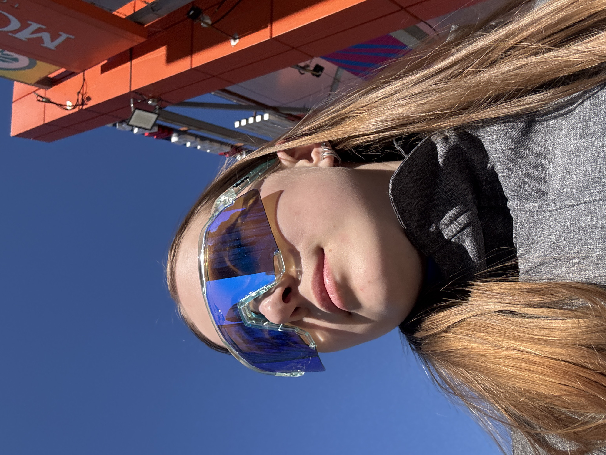 The image shows a person with long, light brown hair, wearing blue-tinted ski goggles. The person's face is angled towards the camera. The background consists of a bright blue sky and an orange-colored structure with visible lighting fixtures. The location is the city of Usolye, Russia.