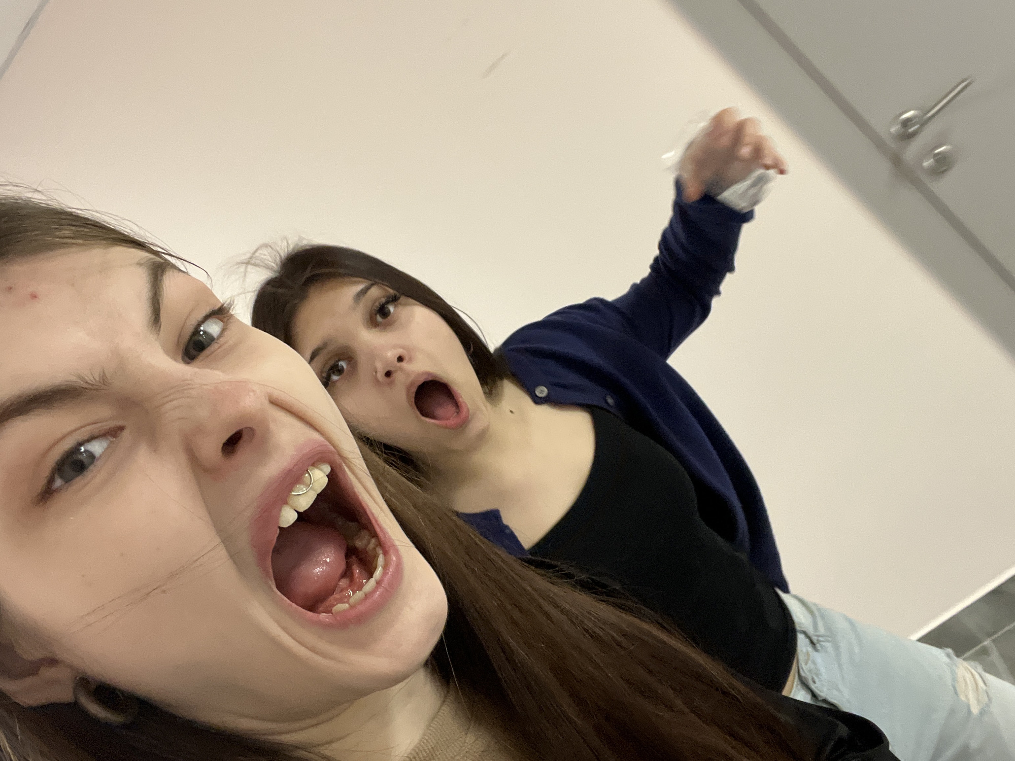 Two young women are captured in a low-angle, close-up selfie. The woman in the foreground contorts her face with a wide, open mouth revealing a piercing on her tooth. The woman in the background, also with an open mouth, is holding a white object aloft. The setting appears to be an indoor bathroom with a white wall and gray fixtures visible in the upper right corner. There is no indication this picture was taken in Saint Petersburg, Russia.